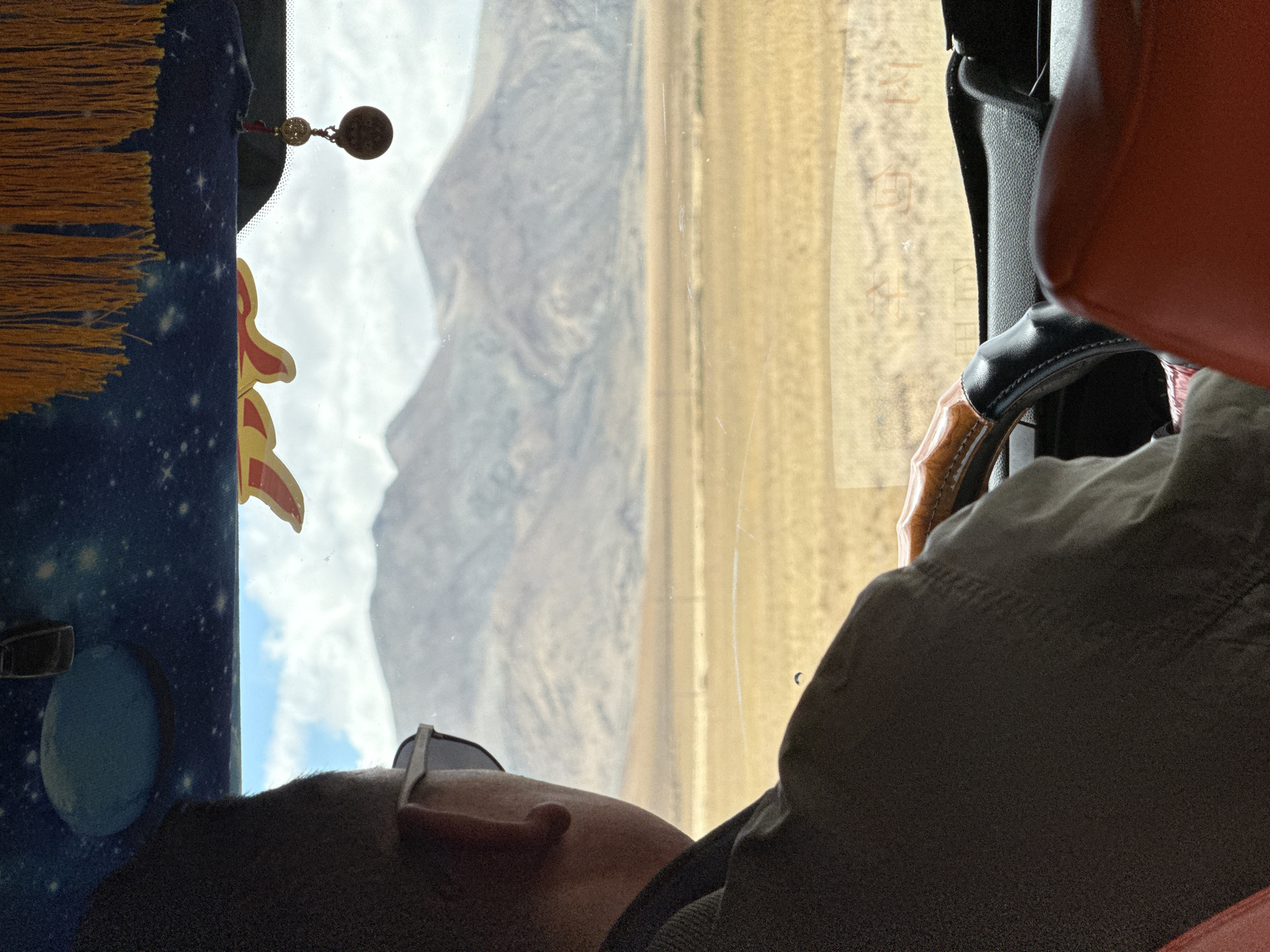A passenger in a vehicle is seen from the shoulders up, wearing sunglasses and looking out the window. Through the window, a mountain range is visible to the left, and a landscape of light brown vegetation is seen to the right. The interior of the vehicle includes a steering wheel, a red seat, and a decorative panel with a space-themed design. The location is likely Kela Township, China, based on the mountainous terrain visible through the window. The passenger is passively observing the scenery.