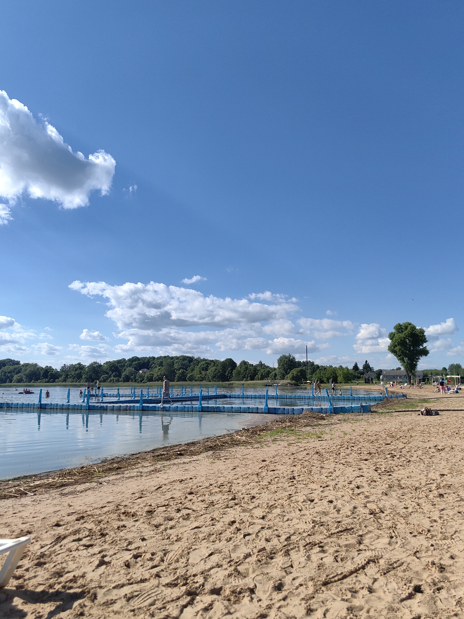 The image depicts a sunny day at a beach on a lake in Myadzyel, Belarus. The foreground is dominated by a sandy beach with footprints visible. In the middle ground, a bright blue floating platform extends into the calm lake, with several people walking on it and in the water. A red inflatable raft is also visible on the lake in the distance. The background features a dense line of green trees and some buildings. The sky is a clear blue with scattered white clouds. The overall scene conveys a sense of leisure and recreation.