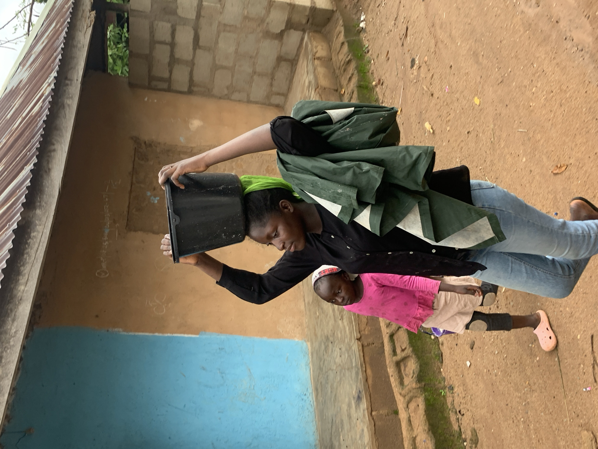 In Barakin Lerawi, Nigeria, a woman walks on a dirt path carrying a black bucket balanced on her head, with a green cloth draped over it. She wears a black long-sleeved shirt and blue jeans. A young girl walks beside her, wearing a pink long-sleeved shirt and a light-colored skirt with dark leggings underneath, and pink Croc-style shoes. Behind them is a building with a blue and brown painted wall, and a corrugated metal roof. The environment is rural, with scattered debris on the ground.