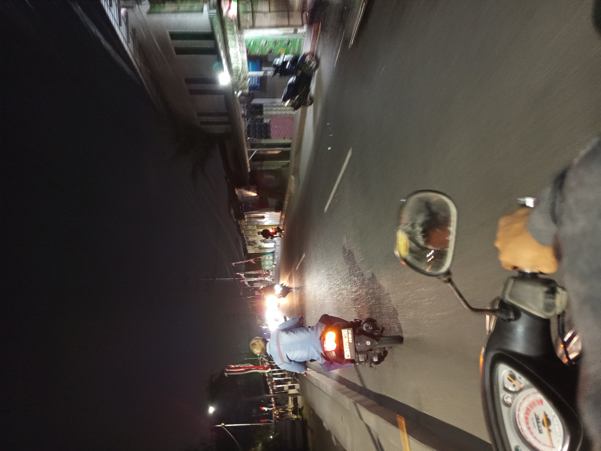The image depicts a nighttime street scene in Tebel, Indonesia, captured from the perspective of someone riding a motorcycle. The foreground shows the handlebars and a portion of the dashboard of the motorcycle the viewer is on, including a speedometer with a red needle. A right hand is visible gripping a throttle.
In the mid-ground, another person is riding a dark-colored scooter ahead of the viewer, facing away. This person is wearing a light blue jacket and dark pants. The scooter has a visible license plate. Further down the road, other motorcycles are visible, some with illuminated headlights, contributing to the nighttime ambiance.
The environment includes a paved road with white lane markings. On the left side of the road, a sidewalk is present, with a street light and several flags visible. On the right side, buildings with illuminated windows line the street, suggesting a commercial or residential area. The overall lighting is dim, characteristic of nighttime, with artificial lights from buildings and vehicles being the primary light sources. The road surface appears wet in places, reflecting the lights.
