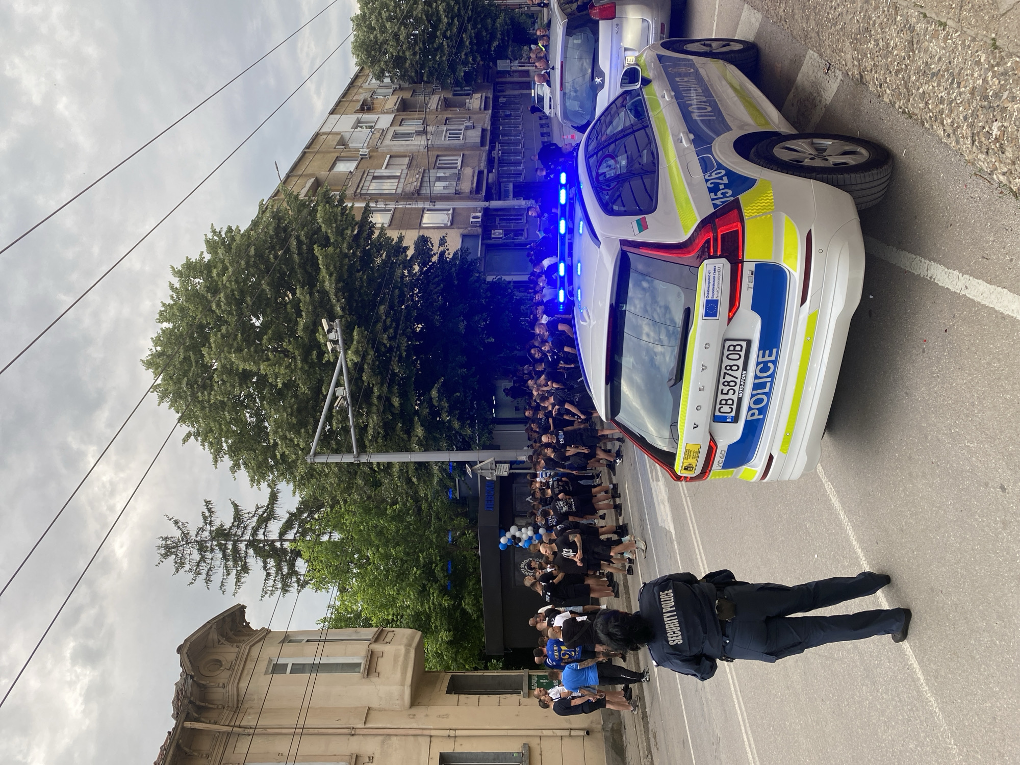 The image captures a scene in Pleven, Bulgaria, featuring a gathering of people in front of a building. Two police cars, with their blue lights flashing, are positioned nearby. A security police officer stands in the foreground, facing the crowd. The majority of the people appear to be wearing black clothing. The license plate on the police vehicle reads "CB 5878 OB" and the car is a Volvo. The building in the background has a black storefront with blue and white balloons. There are large trees in the vicinity and an older building structure to the left. The overall atmosphere seems to involve a regulated or monitored event.