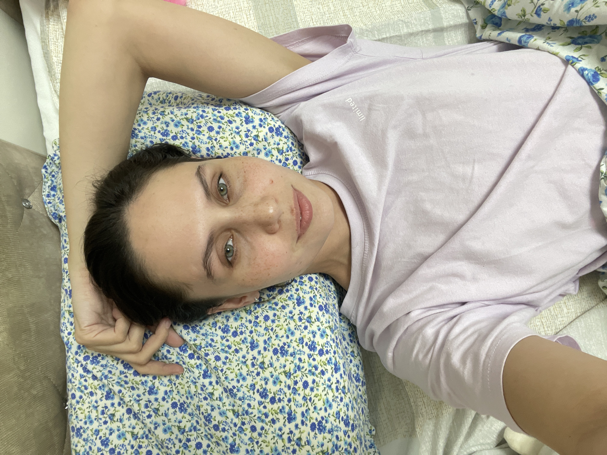 A woman is lying on a bed or sofa. She is wearing a light purple t-shirt and is resting her head on a floral-patterned pillow. Her arm is raised above her head. The environment appears to be a domestic setting. The location is not discernible from the image.