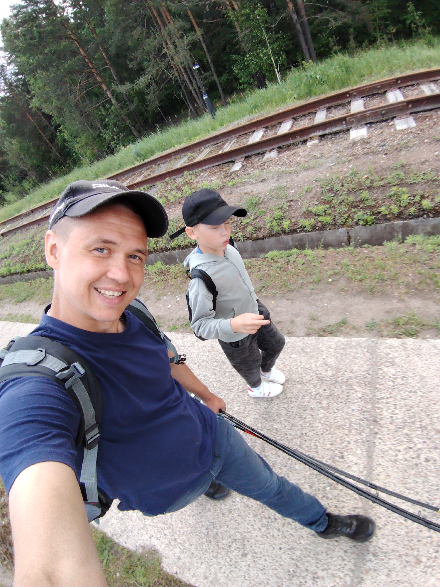 A man and a child stand near railway tracks. The man is taking a selfie. He has on a dark baseball cap, a dark blue t-shirt, blue jeans, and black shoes. He has a backpack on. He is holding fishing rods. The child has on a dark baseball cap, a grey hooded sweatshirt, dark pants, and white tennis shoes. The location is likely near the city of Chaikovsky, Russia based on the surrounding trees and the appearance of the railway tracks.