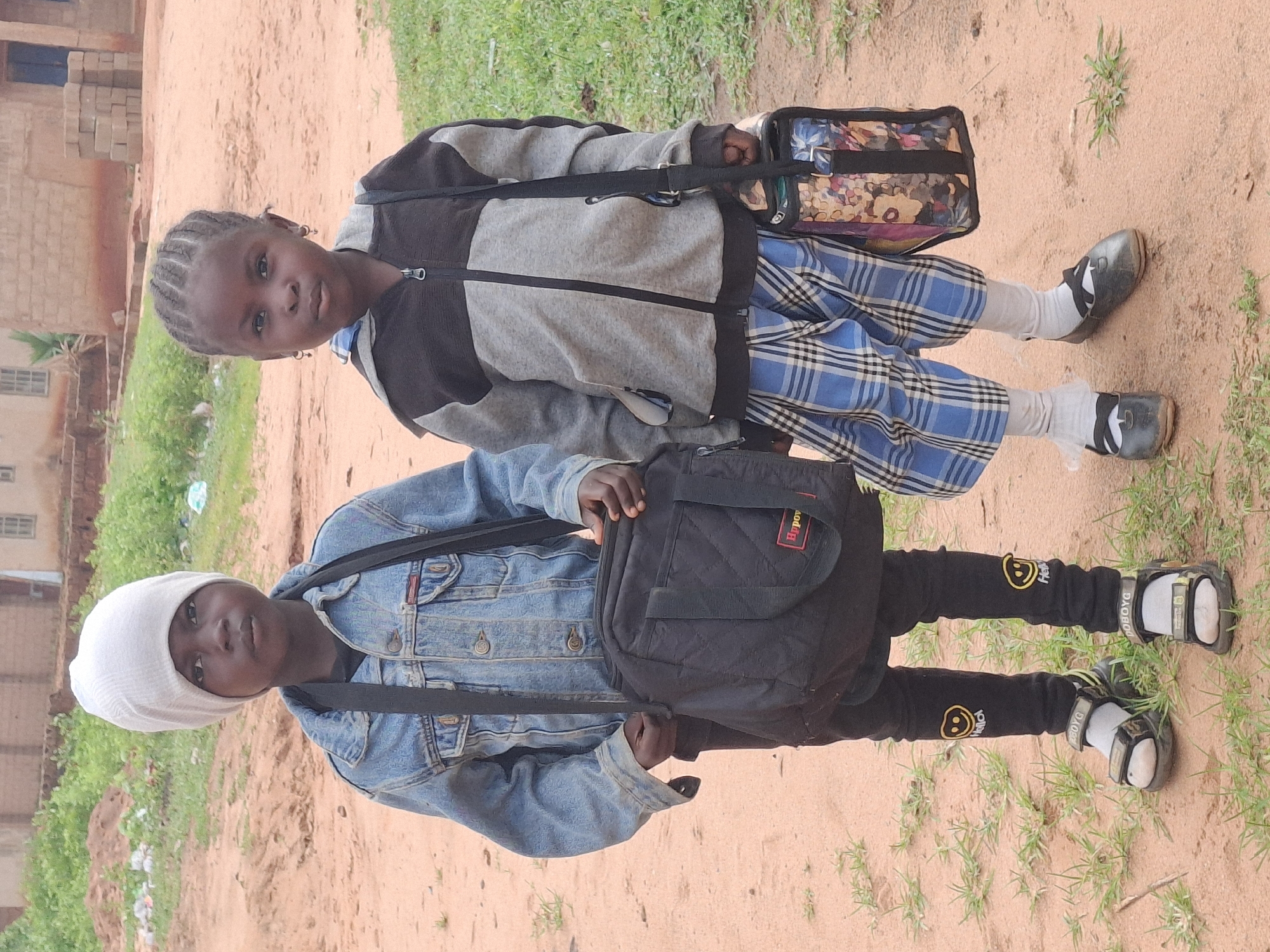 Two young children, a boy and a girl, stand on a dirt path in what appears to be a residential area in Tumfure, Nigeria. The boy, on the left, wears a white beanie, a denim jacket over a black t-shirt, and black pants with smiley face patches. He has a black bag slung across his chest and wears sandals. The girl, on the right, has braided hair and wears a grey and black hooded jacket over a plaid skirt. She carries a colorful patterned bag and wears black shoes with white socks. The background shows simple buildings, some with brickwork, and sparse vegetation. The ground is reddish-brown dirt. The lighting suggests an overcast day.