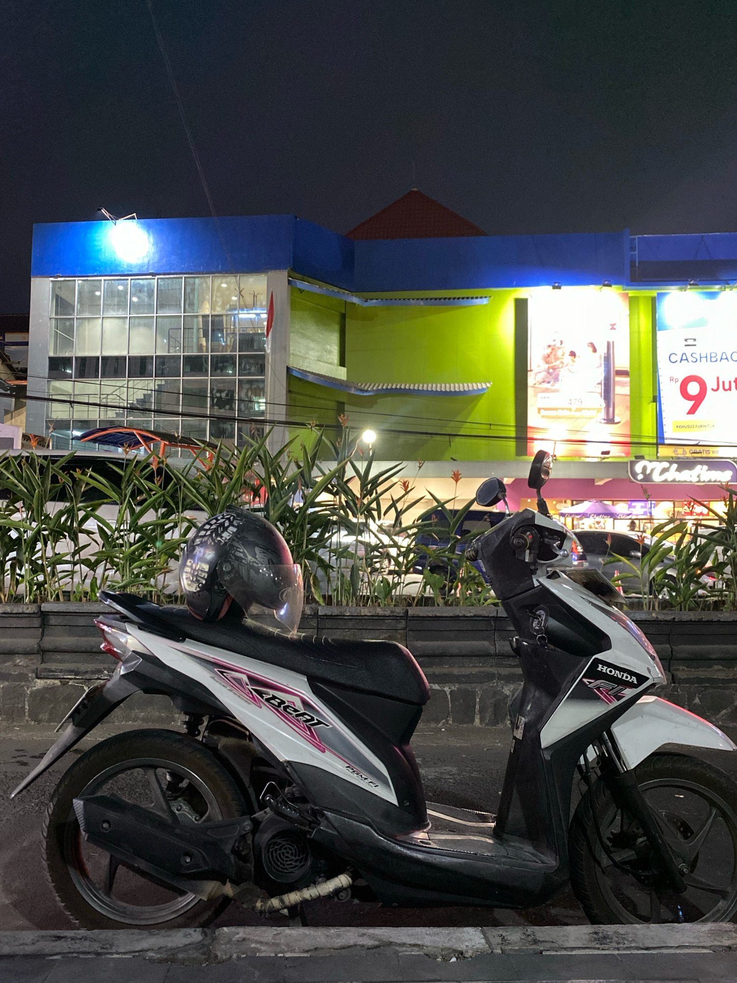 A black and white Honda Beat scooter is parked on a street at night, in front of a brightly lit building. A black helmet with a clear visor rests on the scooter's seat. The building appears to be a commercial establishment, possibly a store or a mall, with large windows and illuminated signage. The scooter is positioned in the foreground, occupying a significant portion of the frame. The background shows the facade of the building, with vibrant colors and advertisements. The lighting suggests it is evening or night. The scene is likely a public area within the city of Magelang, Indonesia, given the context.