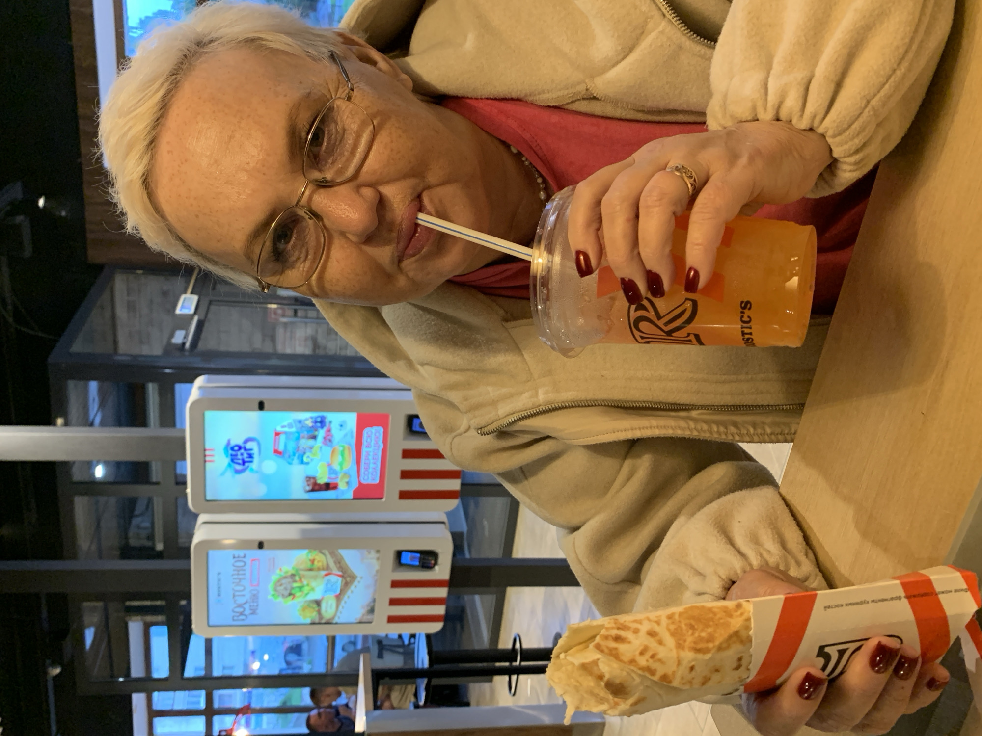 The image captures an elderly woman inside a Rostic's fast-food restaurant, located in Chaikovsky, Russia. The woman, with short, bleached blonde hair, is wearing gold-rimmed glasses and a light beige, textured fleece jacket over a pink top, revealing a pearl necklace. She has dark red nail polish on her fingernails and a gold ring on her right ring finger. She is looking directly at the camera with a slight smile while sipping an orange beverage from a clear plastic cup with a white and blue striped straw, held in her right hand. Her left hand holds a half-eaten wrap or shawarma, partially encased in white paper with red vertical stripes and visible Cyrillic text.
In the background, two digital menu screens are prominent. The screen on the left displays "ВОСТОЧНОЕ МЕНЮ" (Eastern Menu) alongside images of food items. The screen on the right features a "ДЕТО ТИМ" (Deto Team/Kids Team) logo, showcasing a children's meal with a burger, fries, drink, and toys. Both screens are mounted on white structures with red vertical stripes below them, each containing a slot with a dark blue flip phone inserted. Large windows behind the menu screens offer a view of an outdoor urban environment with other blurred figures. The woman is seated at a light-colored wooden table. The interior features dark ceilings and structural elements.
