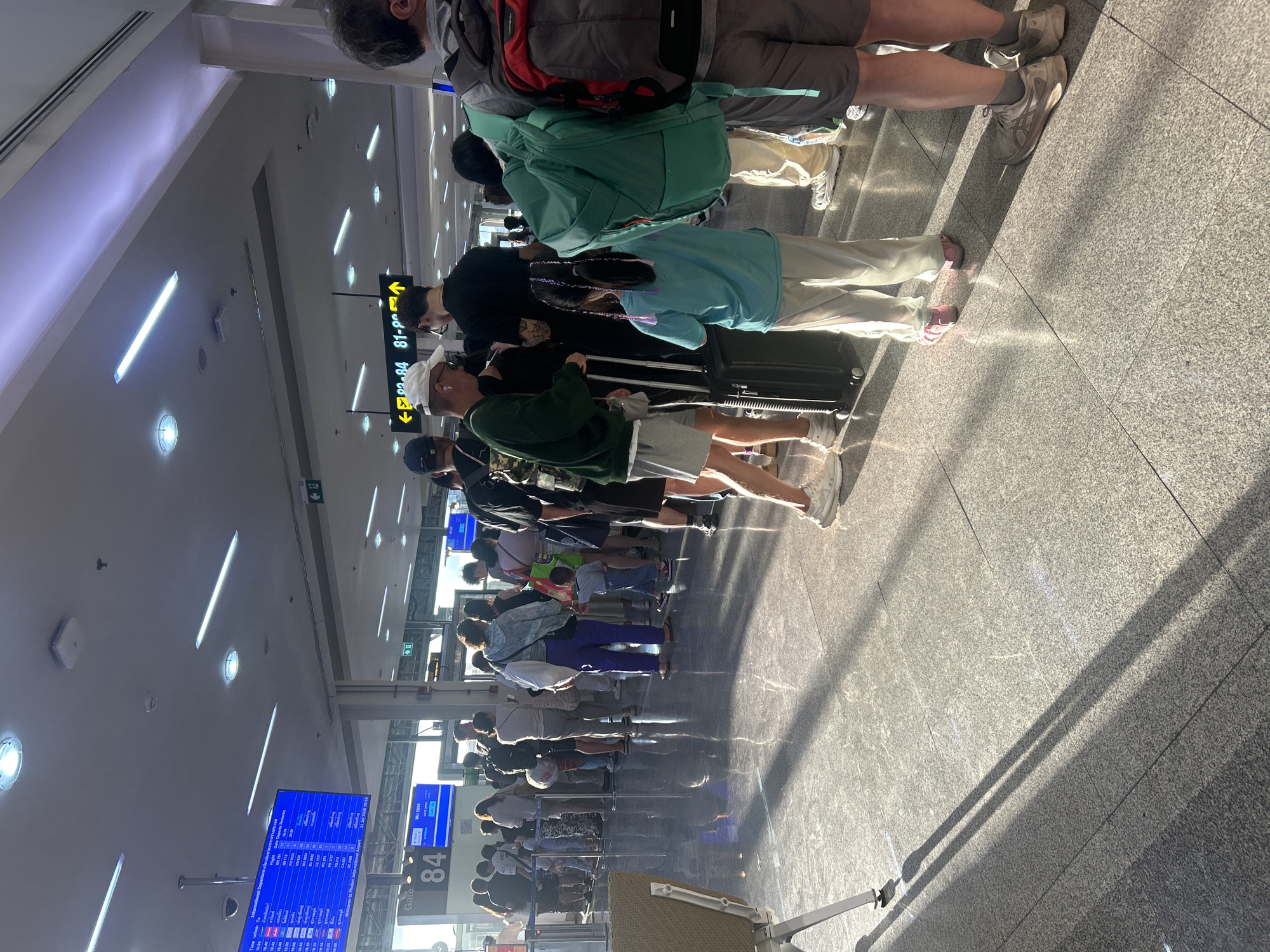 The image depicts a queue of people at an airport in Mai Khao, Thailand. The people are standing in line, with some holding luggage. A digital display board shows flight information, and a gate number "84" is visible. The environment appears to be an indoor airport terminal with tiled flooring and overhead lighting. Sunlight is casting long shadows across the floor, indicating it is daytime. Some individuals are wearing casual attire, and children are present in the queue. The overall scene suggests a typical airport departure or boarding process.