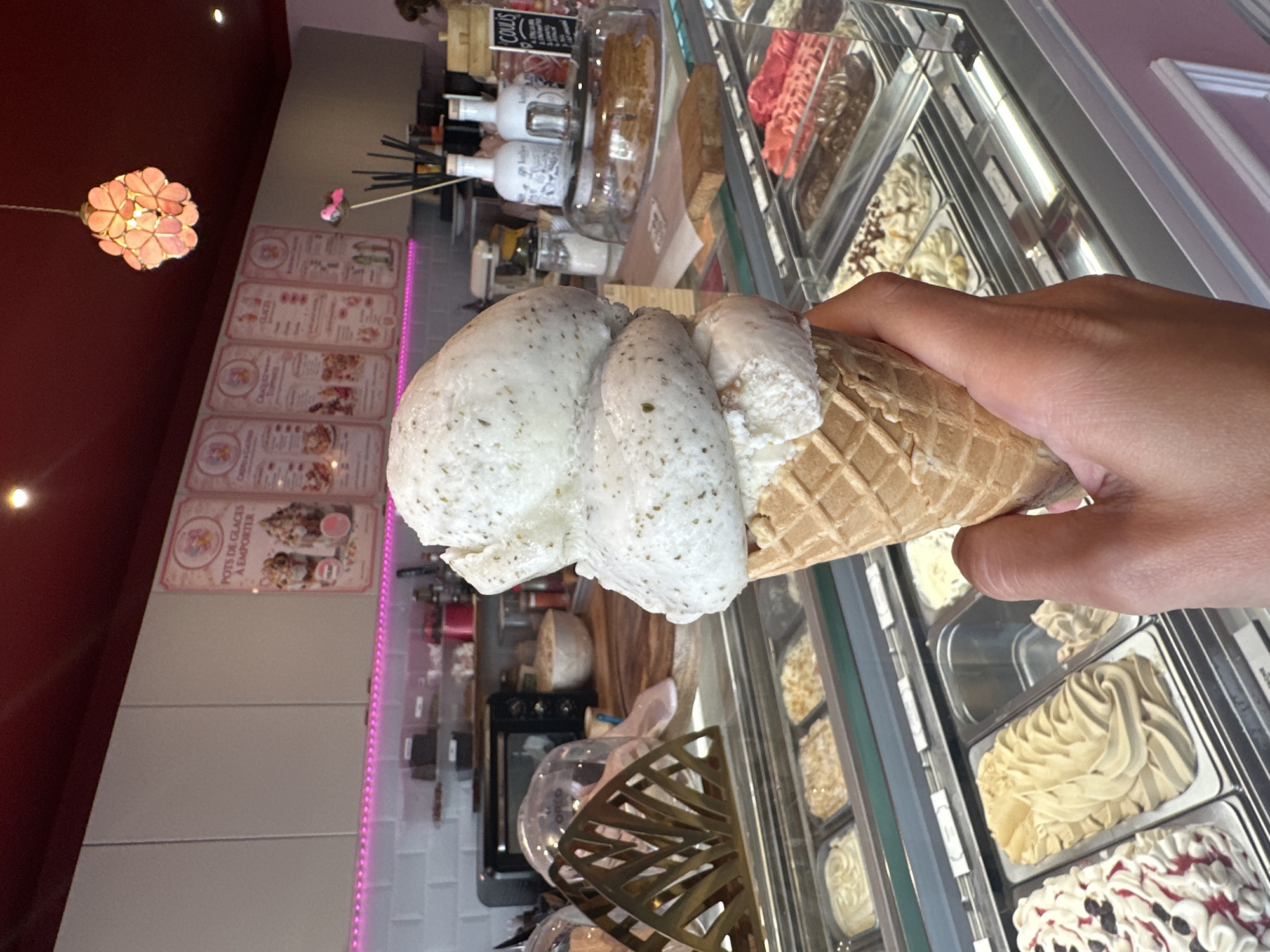A person's hand holds a waffle cone filled with two scoops of white ice cream speckled with dark green herbs, likely a mint or basil flavor. The ice cream is positioned in the foreground, obscuring part of the scene. In the background, an ice cream shop displays various flavors of gelato in a refrigerated case. The shop has a modern aesthetic with white tiled walls, a pink illuminated accent light, and a dark red ceiling. Menus with illustrations of ice cream desserts are mounted on the wall. Behind the counter, bottles of sauces and other condiments are visible. The location appears to be an ice cream parlor in Dieppe, France, suggested by the French text on the menus.