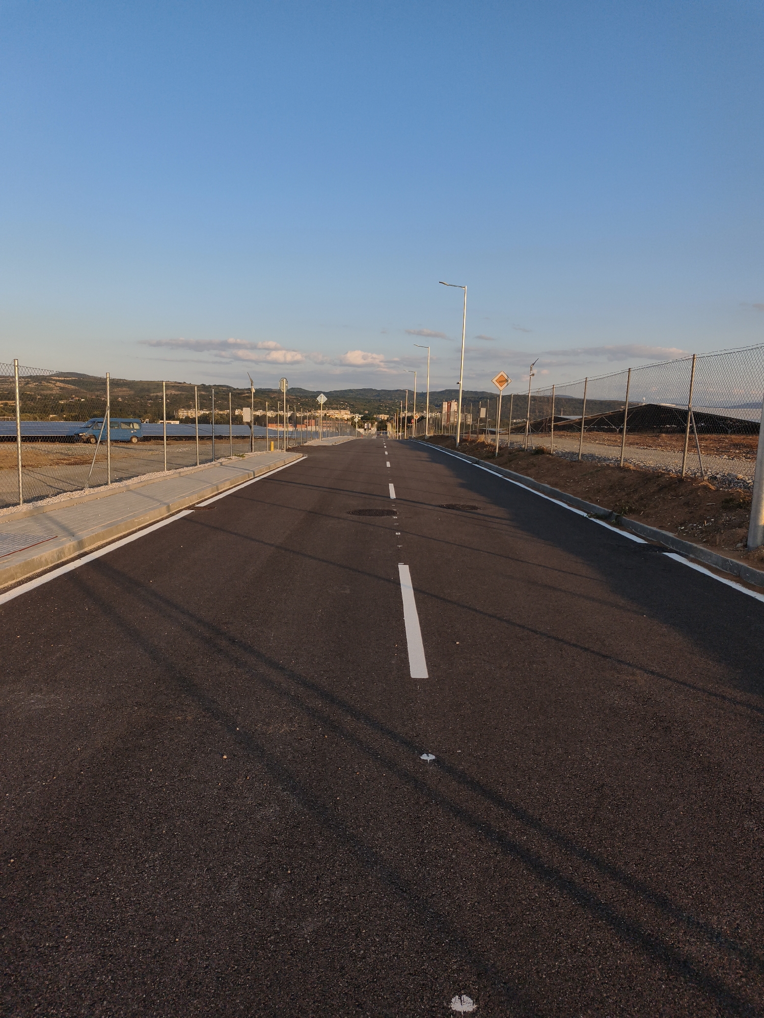 The image depicts a straight, paved road with white lane markings, leading towards a distant horizon. The road is bordered by a chain-link fence on either side, with a sidewalk along the left. Beyond the fence on the left, a solar panel farm is visible, with a blue van parked nearby. On the right side of the road, construction or undeveloped land is present, with a signpost and streetlights along the fence. The sky above is clear blue with a few scattered clouds, and the setting sun casts long shadows across the road. The landscape in the background appears to be hilly and vegetated, with some buildings visible in the distance. There are no people or animals in the image. This scene is likely located on the outskirts of Lovech, Bulgaria, possibly near an industrial area or a newly developed zone given the presence of solar panels and the overall undeveloped nature of the surrounding land.