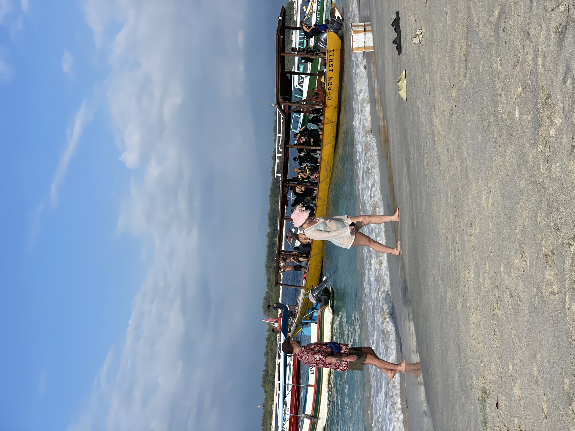 In Unknown, Indonesia, a bright, sunny day unfolds on a sandy beach bordering turquoise waters. Several long, narrow boats, likely for transportation or diving tours, are anchored offshore. The prominent boat in the foreground is a bright yellow vessel with a covered canopy, carrying a group of people, many of whom appear to be divers with scuba gear. The boat is named "O-REN ISHII." To the left of the yellow boat, other boats with colorful stripes are docked.
On the beach, two individuals are walking along the waterline. A man wearing a patterned short-sleeved shirt, shorts, and a blue fanny pack is on the left. Beside him, a woman in a light-colored sheer cover-up and a pink wide-brimmed hat is walking towards the right. Their feet are near the gentle waves lapping at the shore. In the sand near the water's edge, several pairs of sandals and a white box with tape are visible. The background shows a distant green coastline under a partly cloudy blue sky.