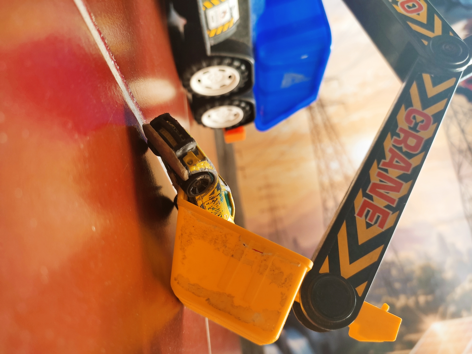 A toy crane, with "CRANE" printed on its arm, is positioned over a yellow toy car. The crane's yellow bucket is scooping up the car. In the background, a toy dump truck with a blue bed is visible. The scene appears to be set on a floor, possibly in a child's room or playroom. There is no discernible connection to the city of Ponorogo, Indonesia, as no specific landmarks or contextual clues indicate this location.