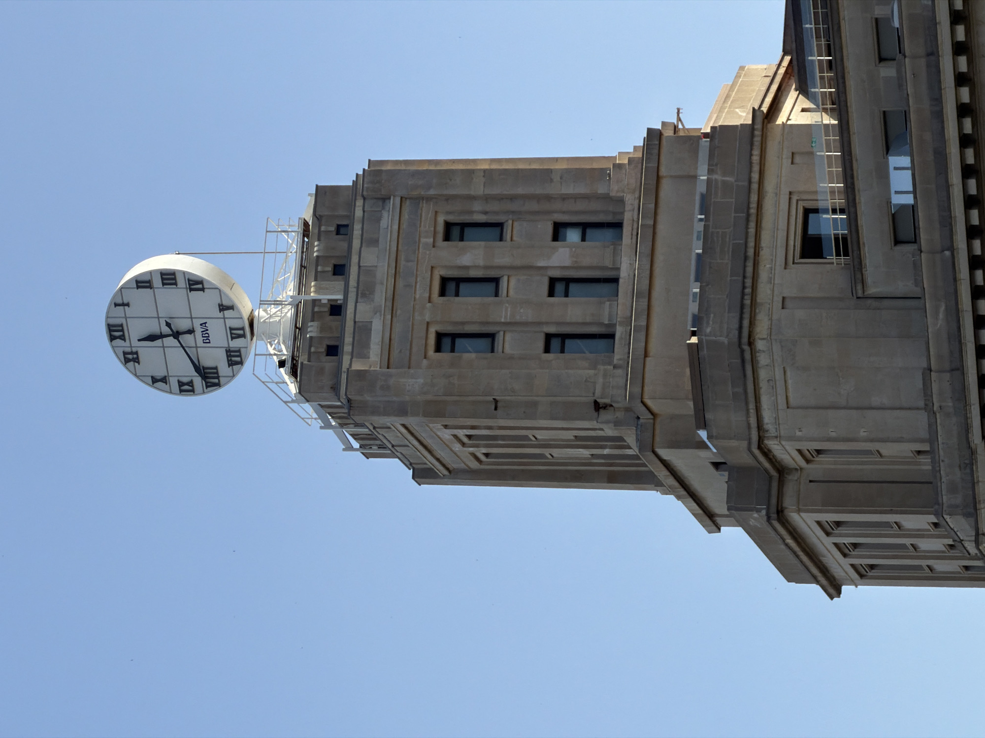 A wide shot captures the upper portion of the BBVA building in Barcelona, Spain, under a clear, pale blue sky. The building, constructed from light brown stone, features a prominent clock tower at its apex. The clock face, round and white with Roman numerals, displays the time as approximately 9:45. The clock is supported by a white, lattice-like structure. Below the clock, the tower's facade is characterized by vertical architectural elements and rectangular windows. The lower sections of the building visible in the frame also exhibit classical architectural styles with repeating window patterns. A railing system is visible on some of the upper ledges. The scene is devoid of people or any visible activity, presenting a static architectural study.