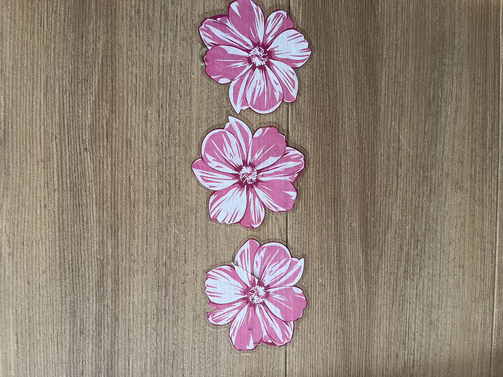 Three pink and white paper flowers are arranged in a horizontal line on a light brown wooden surface. The flowers are the same size and design, with five petals each, and a central stamen area. They are positioned evenly spaced apart. The wooden surface has a visible wood grain pattern running horizontally.