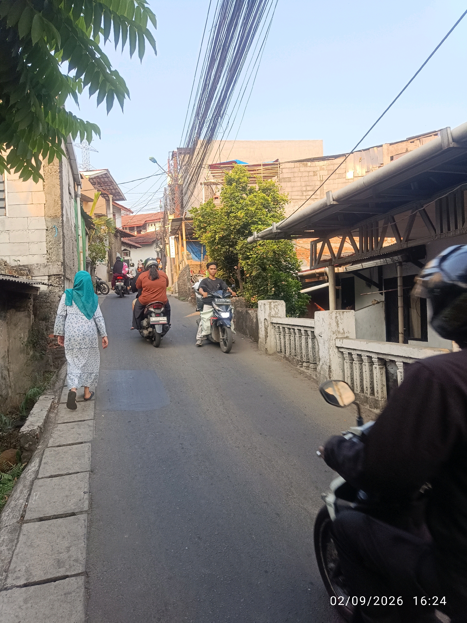 In an outdoor urban scene, a person wearing a long patterned dress and a turquoise hijab walks away from the camera on the left side of a paved road. The person wears flip-flops. The road is lined with a concrete sidewalk and a low concrete barrier with decorative pillars. Several motorcycles are present on the road. One motorcycle in the foreground is ridden by a person wearing a black helmet and dark clothing, with the back of their head and shoulders visible. Further up the road, a motorcycle is ridden by a person in a black shirt, and another motorcycle has a person in an orange shirt as the rider. More motorcycles and people are visible in the background. Buildings, including houses and shops, line both sides of the narrow street. Overhanging power lines and greenery from trees are also part of the scene. The lighting suggests it is daytime. A timestamp in the lower right corner reads "02/09/2026 16:24".