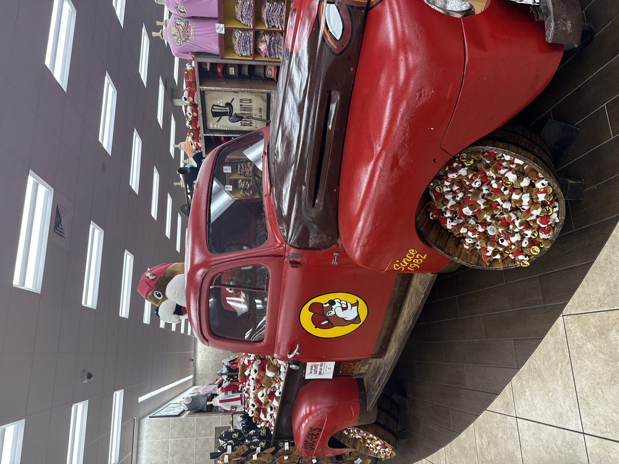 The image displays an indoor scene within a Buc-ee's travel center, located in Fort Valley, United States. The primary subject is a red, vintage-style pickup truck functioning as a merchandise display. The truck features a dark brown, distressed finish on its hood and upper fenders. A large yellow circular logo of Buc-ee the beaver wearing a red cap is affixed to the driver's side door, and "Since 1982" is painted in yellow on the lower front fender. The truck bed is filled with numerous small, identical Buc-ee beaver plush toys. The vehicle's wheels are replaced with wooden barrels, also overflowing with the same plush toys. A large, oversized plush head of Buc-ee, adorned with a red cap, is positioned above the truck's cabin.
The immediate environment consists of a tiled floor, transitioning from lighter square tiles to darker rectangular planks where the truck is situated. The ceiling is a white suspended grid with multiple rectangular fluorescent light fixtures providing overhead illumination. A small black dome security camera is visible on the ceiling. In the background, retail display shelves are visible, stocked with Buc-ee's branded merchandise including pink t-shirts displayed on mannequins and folded, as well as hats and other apparel. A sign for "Beaver Hat Co Est. 1860" with a top hat logo is positioned behind the truck's hood. A white sign attached to the truck's bed warns, "FOR YOUR SAFETY DO NOT CLIMB ON OR IN DISPLAY." No people are present in the scene.