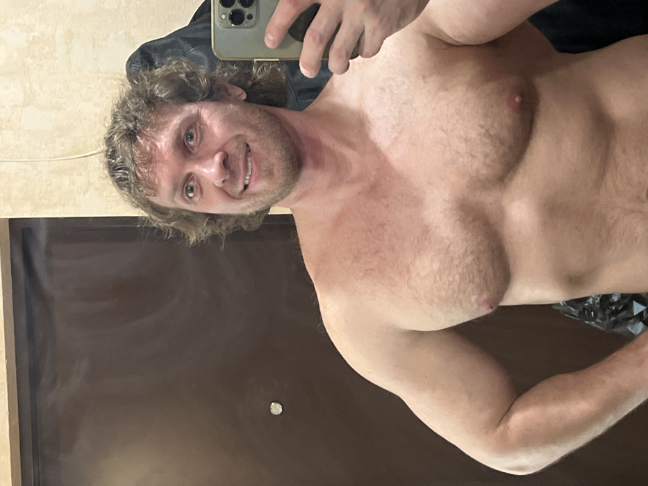 The image displays an adult male taking a mirror selfie indoors, within what appears to be a private residential setting in Saint Petersburg, Russia. The subject is a light-skinned male, approximately in his 30s-40s, with curly light brown hair and a short beard. He is shirtless, revealing a muscular physique with moderate chest hair, and is smiling while looking directly at the camera. His right hand holds a smartphone, identifiable by its multi-lens camera array, angled to capture his reflection. The background on the left consists of a dark brown panel door featuring a circular silver-colored peephole or knob. The wall to the right of the door and behind the subject exhibits a light, textured finish. A dark garment is visible hanging on the wall behind the subject's right shoulder, and a crumpled dark plastic bag is partially visible in the lower-left background. No distinct architectural features are present to provide a more precise location within the city.
