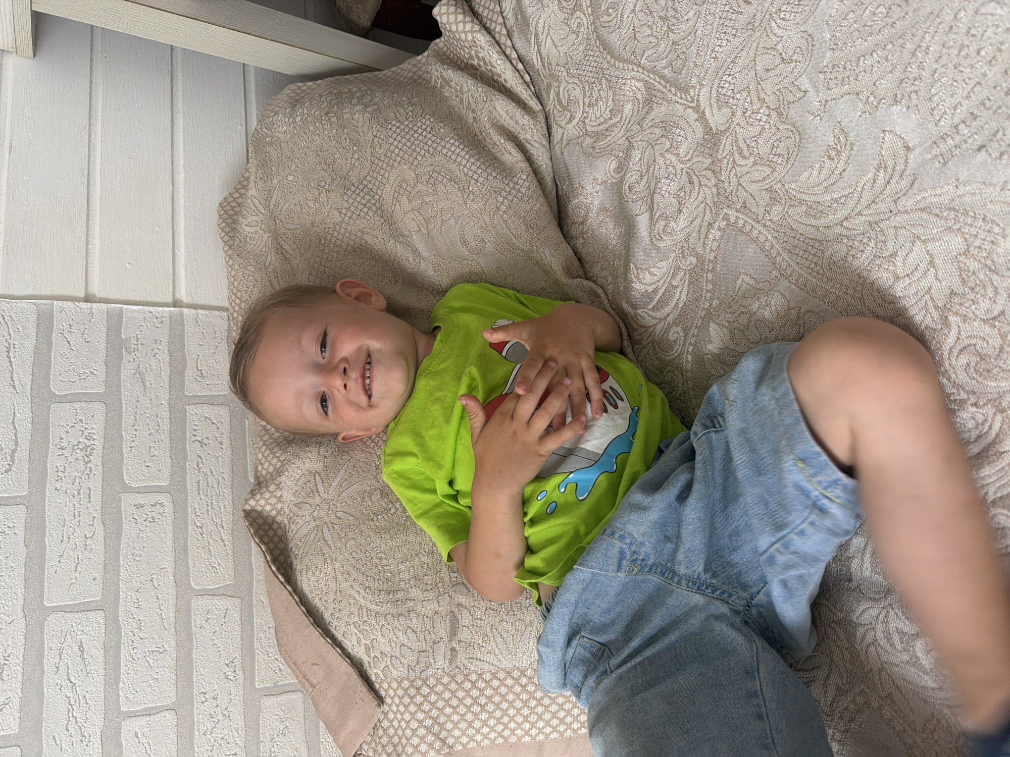 A young boy is lying on his back on a bed in an indoor setting. He is wearing a bright green t-shirt with a graphic print and denim shorts. His hands are clasped over his stomach, and he is smiling at the camera. The bed has a patterned beige duvet and a pink pillow. The background features a white brick wall and a wooden wall. The scene is likely a bedroom. There is no indication that this image was taken in Sochi, Russia.