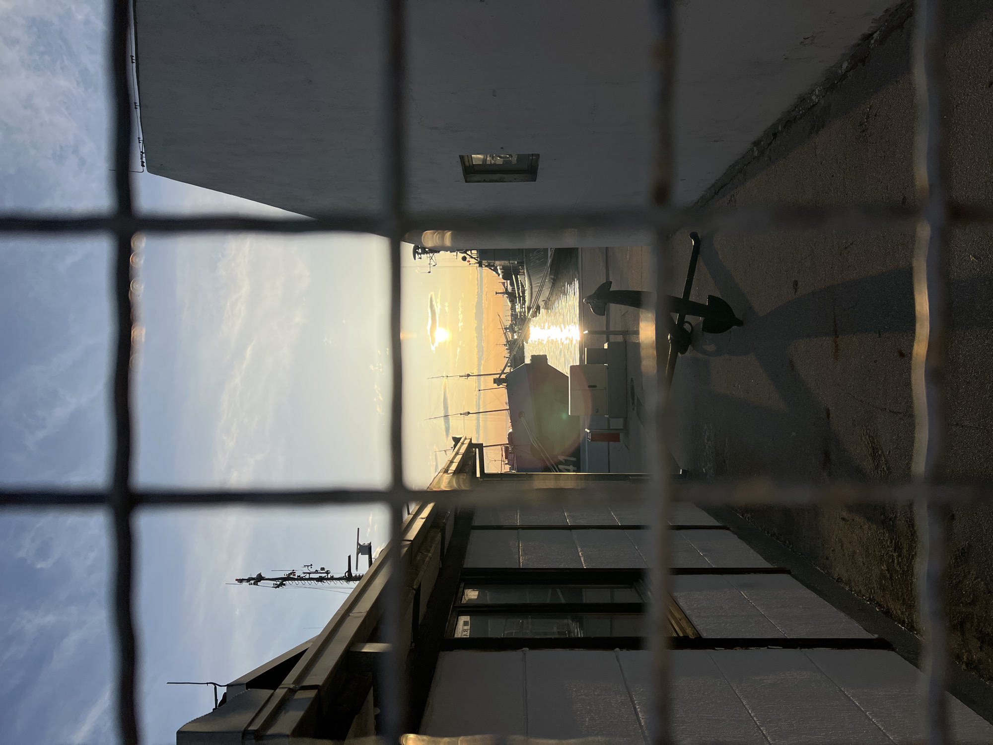 The image, shot from behind a wire fence, presents a horizontally flipped view of the city of Bar, Montenegro, near the harbor. A sunset is visible between buildings, reflecting light off the water. A docked boat is visible in the distance. An anchor is near the camera. The scene is framed by buildings on either side of a walkway, with the setting sun providing backlighting. The sky has blue and white streaky clouds. The ground appears to be asphalt.