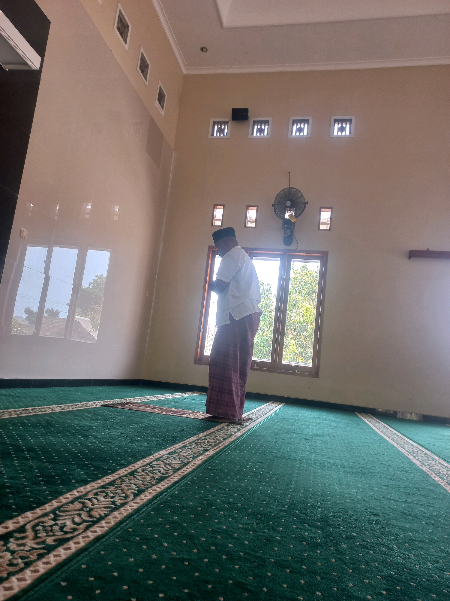 A man wearing a white shirt, a dark cap, and a red and white checkered sarong is standing in prayer on a green carpet within a room. The carpet has decorative patterns. The room has a high ceiling with recessed lighting fixtures and decorative ventilation openings along the upper walls. A ceiling fan is mounted on the wall above a window. The window is divided into two panes and overlooks an outdoor scene with trees and buildings. The walls are painted a light, neutral color. The image appears to have been taken in a mosque or prayer room. The metadata indicates the location as Nguntoronadi, Indonesia.