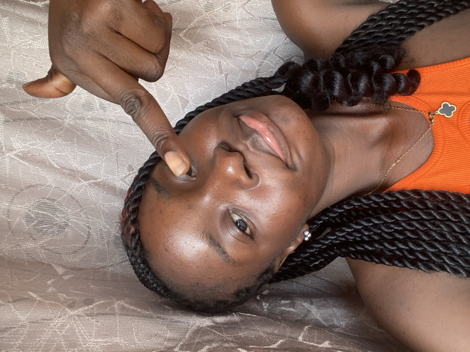 A woman with dark skin and braided hair lies on a patterned surface. She wears an orange top and a gold necklace with a clover pendant. Her hand is raised, with a finger pointing at her eye. Her head is slightly tilted, and she has a gentle expression. The photo's composition is close-up and intimate.