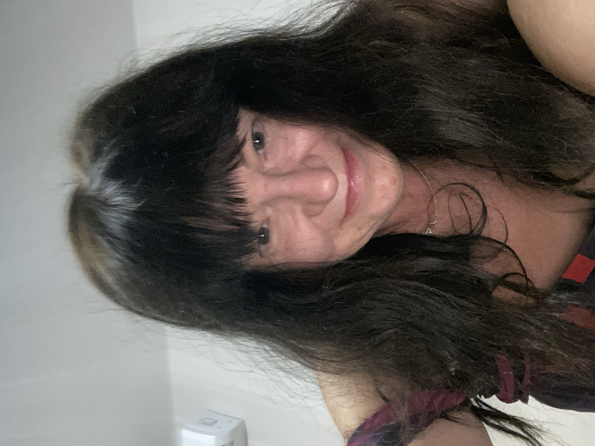 A woman with long, dark hair and bangs poses for a selfie indoors. She is wearing a dark top with a burgundy and red pattern. A white air conditioning unit is visible on the wall behind her. The lighting is soft, casting shadows on her face.