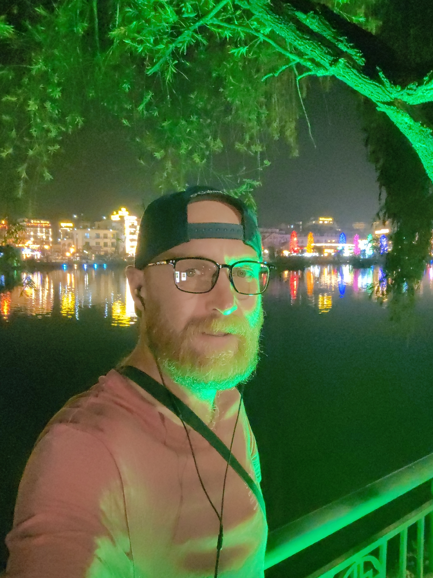 An adult male stands outdoors at night, positioned next to a body of water identified as Sa Pa Lake in Sa Pa, Vietnam. The background includes several illuminated multi-story buildings, one prominently displaying "SAPA HOTEL," which reflect in the dark water. The male subject is depicted from the chest up, looking directly at the camera with a slight smile. He has a beard, wears dark-framed eyeglasses, a dark baseball cap worn backward, and a light-colored short-sleeved t-shirt. A dark strap crosses his right shoulder/chest area, and wired earbuds are visible around his neck. The immediate foreground, including the subject's face and beard, and overhead tree foliage, is strongly illuminated by a green light source. A dark green metal railing is visible in the lower right foreground. The buildings across the water are brightly lit with diverse colors, including yellow, orange, blue, red, and pink, which create prominent, vibrant reflections on the lake's surface.