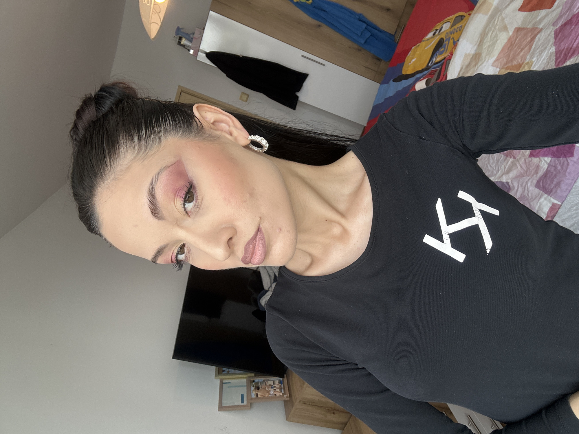 A woman with dark hair pulled back in a bun is positioned in front of a wall with a television. She is wearing a black long-sleeved shirt with a white graphic on the chest. Her makeup includes pink eyeshadow and lipstick. She has a white, circular earring. Behind her is a partially visible bed with a colorful comforter. The background shows a white wardrobe and a wooden dresser with framed photos on top. The location appears to be an interior living space. There is no indication of it being in Burgas, Bulgaria.