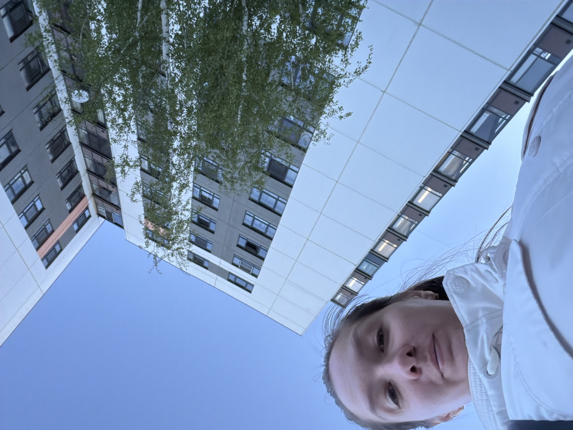 A low-angle shot captures a woman's face in the foreground, with buildings and a tree filling the background. The woman wears a white jacket. The buildings feature light-colored facades, windows, and architectural details. A tree with green leaves is partially visible between the buildings. The sky is clear and blue. The location is likely Unknown, Russia.