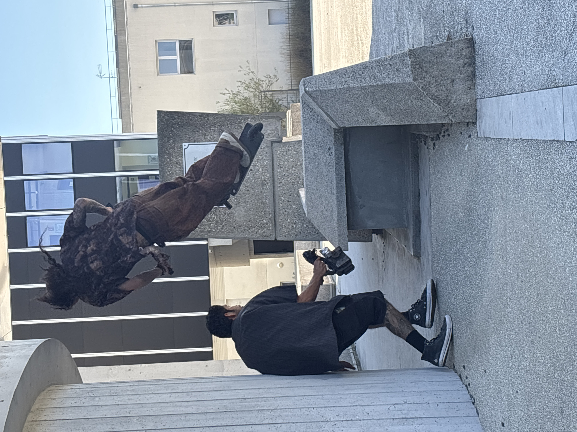 The image captures a dynamic moment in a public outdoor space, likely a plaza or park, with modern architecture in the background. A person with long, dark, braided hair is captured mid-air, appearing to be performing a stunt or jump on what might be a scooter or similar equipment, wearing a patterned shirt and brown pants. Below and to the left, another person, wearing a dark short-sleeved shirt and black shorts, is holding a video camera and looking up towards the airborne individual, seemingly filming them. The ground is paved with a rough texture, and large concrete structures are present, including a curved pillar on the left and angular blocks forming part of the landscape. In the background, a multi-story building with dark vertical elements and windows is visible, along with a lighter-colored building with a visible window and a weather vane on its roof. The sky is clear and blue. The location is specified as Lyon, France.