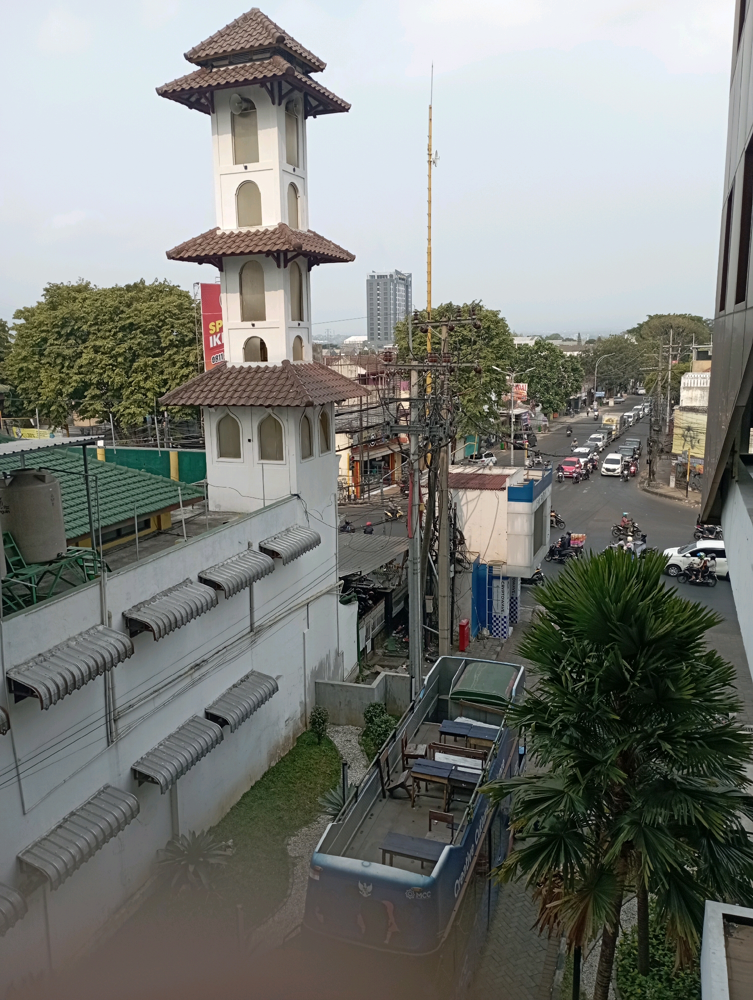 This is an aerial photograph taken from a high vantage point in Malang, Indonesia, looking towards a busy intersection. In the foreground, on the left, is a white minaret with a tiered, brown tiled roof. Adjacent to it is a white wall with multiple grey awnings. Below the wall, a small garden area with a palm tree is visible. To the right of the minaret, a street is teeming with traffic, including cars, motorcycles, and a bus. The buildings lining the street are a mix of modern and older structures, with electrical poles and wires crisscrossing overhead. The sky is overcast.