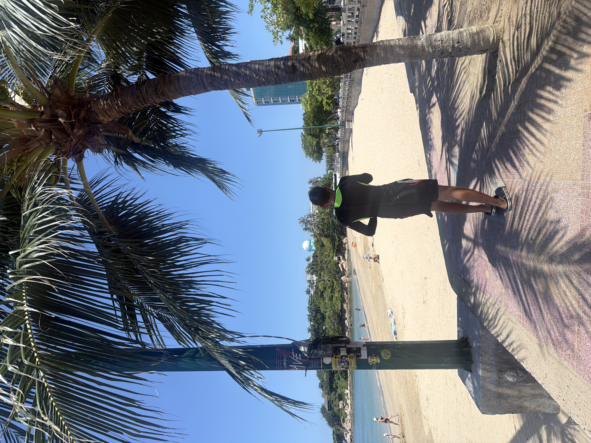 A person, facing away from the camera, stands on a paved walkway near a sandy beach in Nha Trang, Vietnam. The person is wearing a black wetsuit with neon green accents and black shorts over it, along with black and white slides. They are holding a black crossbody bag. The walkway is adorned with patterned tiles and cast with the shadows of palm fronds from trees lining the path.
To the left of the walkway is a sandy beach with a few people scattered about. Some are walking along the shoreline, while others are sitting or lying on the sand. The ocean is visible in the background, with gentle waves lapping at the shore. In the distance, a body of water meets a lush green hillside dotted with buildings. One prominent building, a tall, modern, light blue skyscraper, stands out against the clear blue sky. A street with trees and a sidewalk runs parallel to the beach, with motorbikes and other vehicles visible in the distance. A lamppost stands near the center of the scene.
A tall, thick palm tree trunk is prominently positioned to the right of the walkway, casting a distinct shadow. A metal pole, adorned with various stickers and flyers, stands closer to the beach. The overall scene suggests a warm, sunny day, likely in the late morning or early afternoon given the angle of the sun and the shadows.