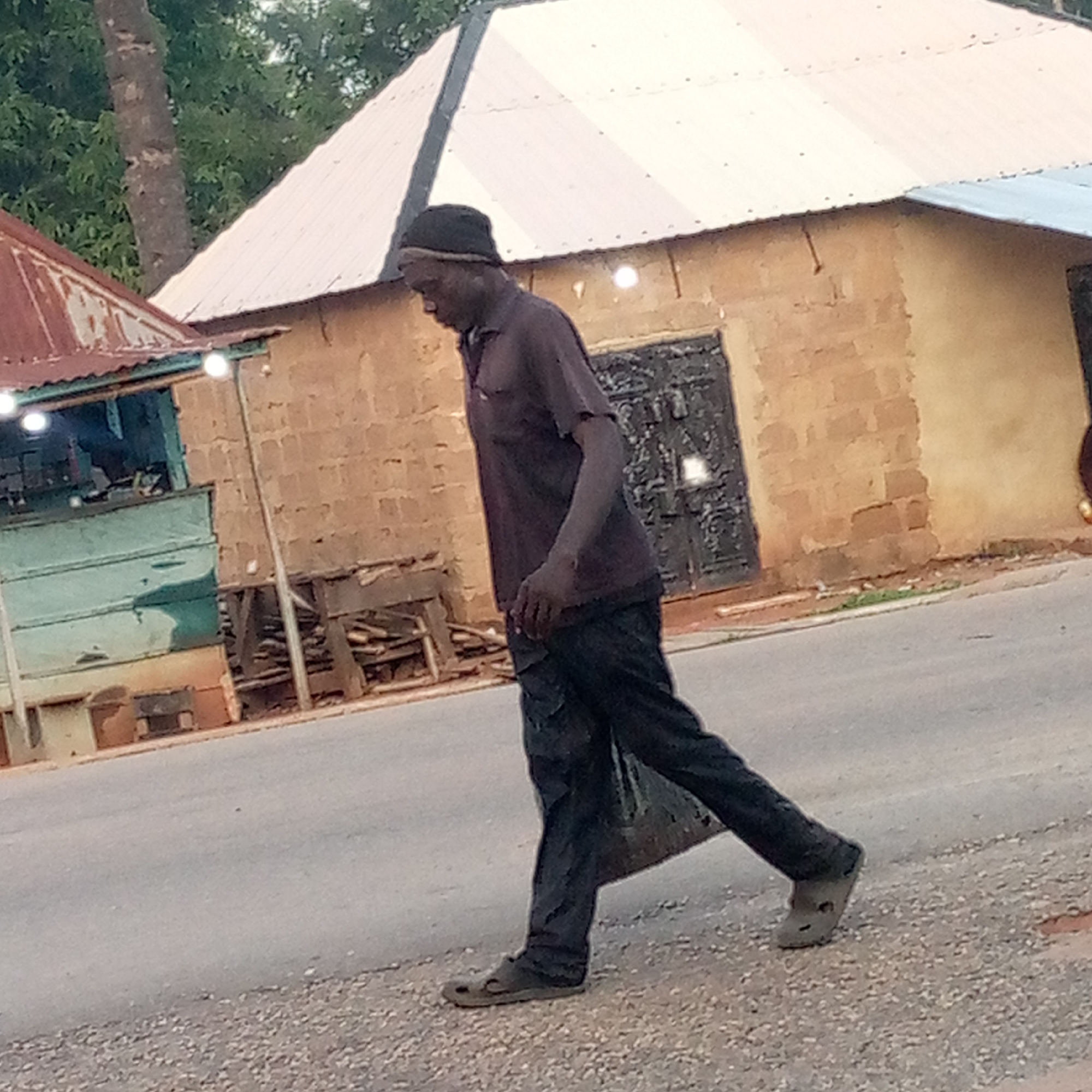 The image depicts a man walking on a paved road in what appears to be a semi-urban or rural setting in Mararaba, Nigeria. The man, with dark skin, is captured in a full-body shot, moving from left to right across the frame. He is dressed in a dark skullcap, a dark short-sleeved polo shirt, dark trousers, and dark slip-on shoes, possibly Crocs-style footwear. He appears to be carrying a dark bag or item in his left hand, which hangs by his side.
In the background, two buildings are visible. To the left, a smaller structure, possibly a market stall or shop, has a rusty reddish-brown corrugated iron roof and teal or light blue painted walls. Several small, bright artificial lights are illuminated along its facade. Behind this, a larger building dominates the mid-ground, featuring a light-colored, likely white or silver, corrugated iron roof and light brown, textured walls, suggesting construction with bricks or mud-bricks. A dark, possibly metallic, door is visible on this larger building. A tree trunk and green foliage are present in the far background on the left side of the image.
The lighting suggests either early morning or late afternoon/evening, indicated by the overall soft light and the active artificial lights. The road appears paved, with a gravelly or dirt shoulder. No visible text is present in the image.