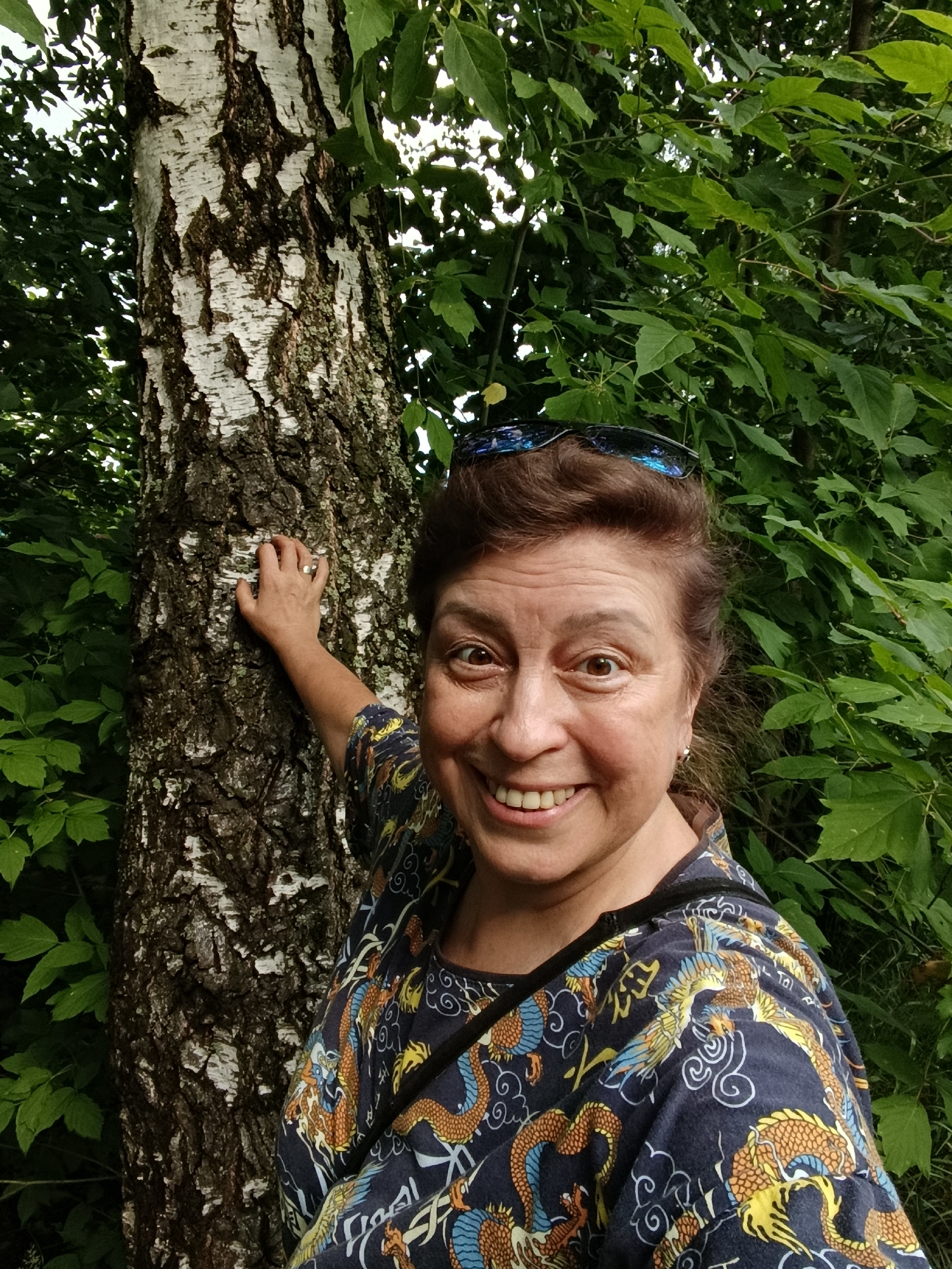 A woman is taking a selfie outdoors, with a birch tree to her left and lush green foliage filling the background. She has brown hair, brown eyes, and is smiling broadly at the camera. She is wearing a dark-colored shirt with a vibrant pattern of golden dragons and blue accents. A silver ring is visible on her left hand, which is resting on the trunk of the birch tree. Sunglasses are perched on top of her head. The setting appears to be a natural environment, possibly a park or wooded area within the city of Vereya, Russia.