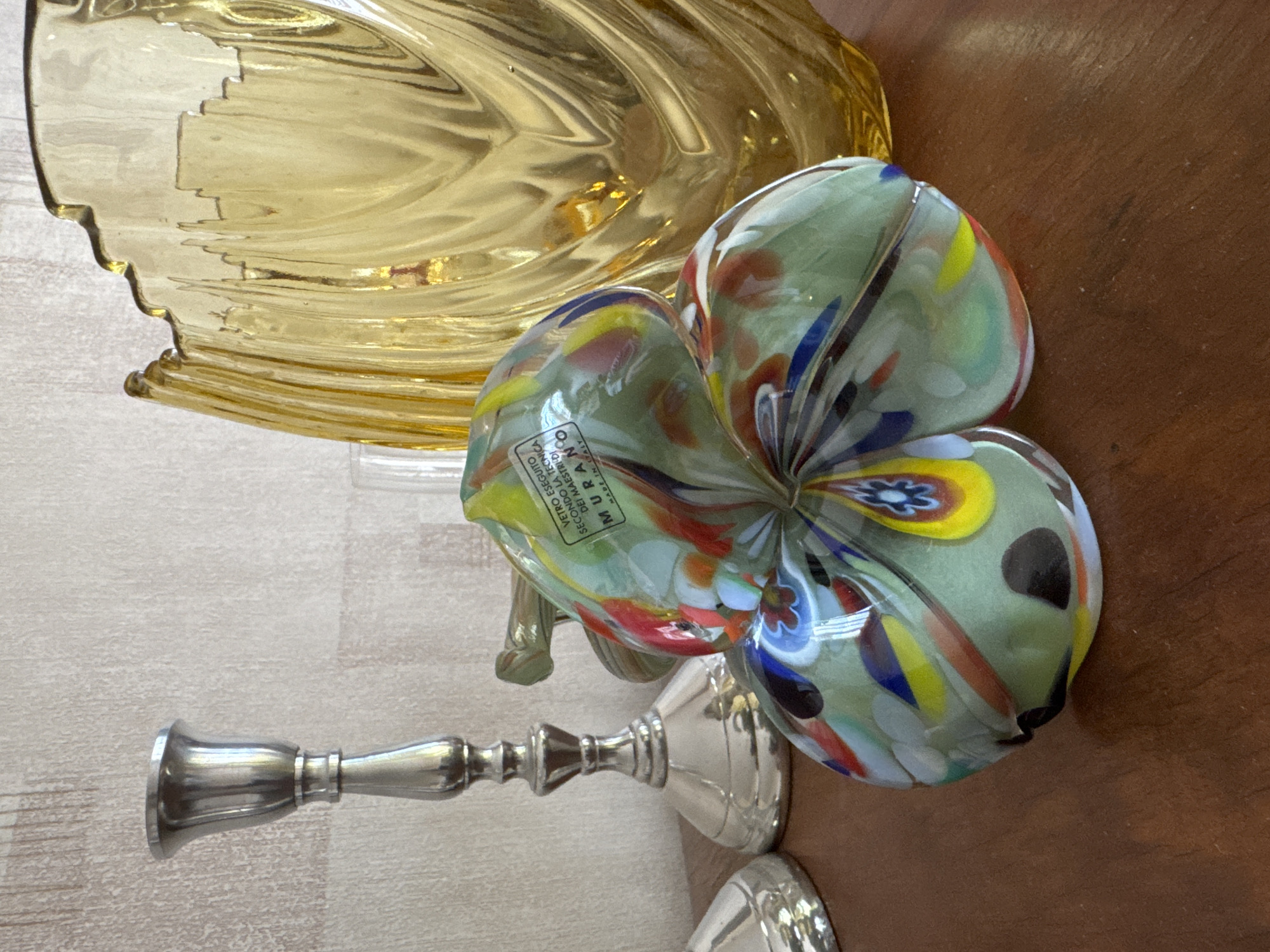 The image shows a tabletop setting with several objects. A golden, ornate glass bowl is partially visible in the upper left. A flower-shaped glass object with a Murano label rests near the bowl. A silver candlestick lies to the left of the flower. The surface the objects rest on appears to be partially wood and partially a lighter, possibly textured surface. The location is not identifiable from the image.