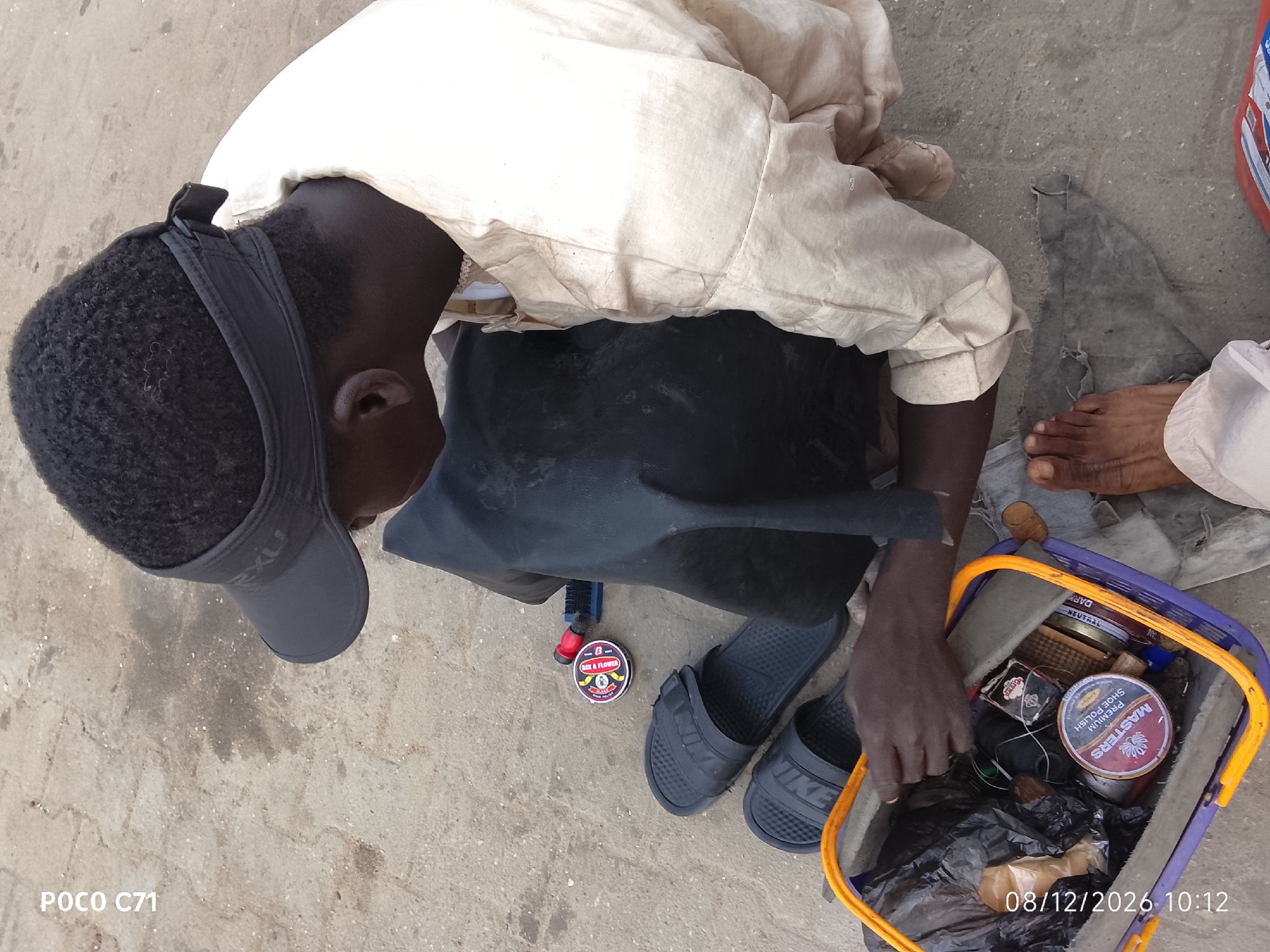 An overhead shot captures a person in Lekki, Nigeria, bent over a shoe-shining kit. The individual wears a light-colored long-sleeved shirt and dark trousers, accessorized with a dark gray visor. They are barefoot, wearing dark slip-on sandals. To their left, a shoe brush and a tin of shoe polish are visible on the ground. To the right, a purple and orange basket, overflowing with shoe-polishing supplies including tins of polish, brushes, and rags, rests on the ground. The scene unfolds on a paved surface, likely a street or sidewalk, with scattered debris. A red plastic container is partially visible in the upper right corner. The image is timestamped "08/12/2026 10:12."