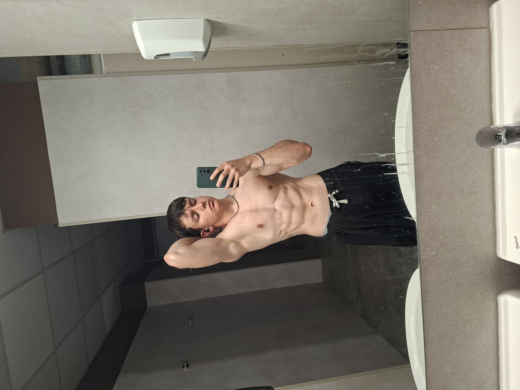 A young man is captured in a mirror selfie, positioned horizontally, possibly in a public restroom or changing room in Plovdiv, Bulgaria. He is shirtless, revealing a toned physique with visible abdominal muscles, and wears black shorts or pants with a white drawstring. His left arm is bent, resting behind his head, while his right hand holds a dark green smartphone, which he uses to take the photo. He has dark hair, a neutral expression with his eyes slightly closed, and is wearing a silver chain necklace, a silver bracelet on his left wrist, and a dark earbud in his left ear.
The setting, viewed through the mirror's reflection, includes light grey tiled walls and darker partitions, suggesting stalls or cubicles. Above the man's reflection, a white paper towel dispenser is visible. To the right of the reflection, part of a white sink with a chrome faucet and a beige speckled countertop are seen. Faint smudges or water droplets are present on the mirror's surface, particularly near the sink area. The word "Yakimas" is faintly visible on the white sink or countertop. The overall lighting is artificial, consistent with an indoor public facility.
