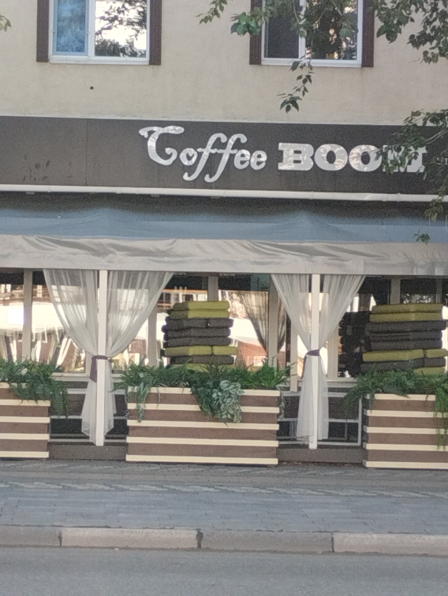 The image shows the exterior of a coffee shop named "Coffee Boom" in Kostanay, Kazakhstan. The coffee shop has a dark brown facade with the name "Coffee Boom" prominently displayed in silver, stylized lettering. Above the facade, there are two windows. The lower portion of the coffee shop features a covered outdoor seating area with white curtains and planters filled with greenery. The planters are made of wood with horizontal stripes in brown and white. In front of the planters, there are stacks of cushions, some of which are green. The ground in front of the coffee shop is paved with interlocking gray bricks. The overall scene suggests a pleasant outdoor seating area for a coffee establishment.