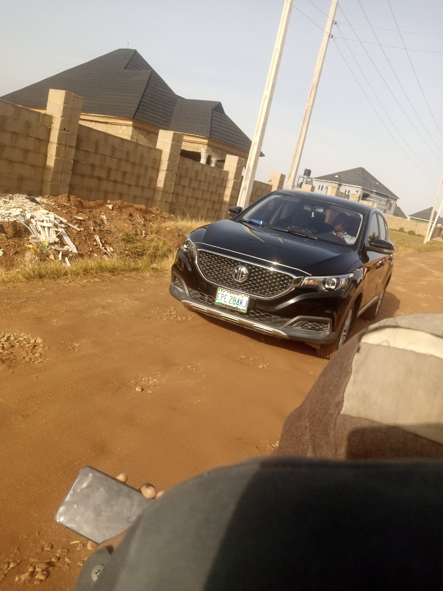 An outdoor, eye-level medium shot captures a black MG car parked on a dirt road in front of a construction site. The car, facing forward with its right side slightly angled away from the camera, has its headlights on. A driver is visible behind the steering wheel, and a license plate reads "LAGOS EPE 284KJ". To the left of the car, a wall constructed of concrete blocks encloses a building under construction, with piles of debris and building materials scattered nearby. The background features a few more houses with dark-tiled roofs and utility poles with electrical wires. In the foreground, a hand holds a smartphone, and a person's lap is visible, suggesting the photo was taken by someone seated or standing close to the ground. The overall scene depicts a developing area under a clear sky.