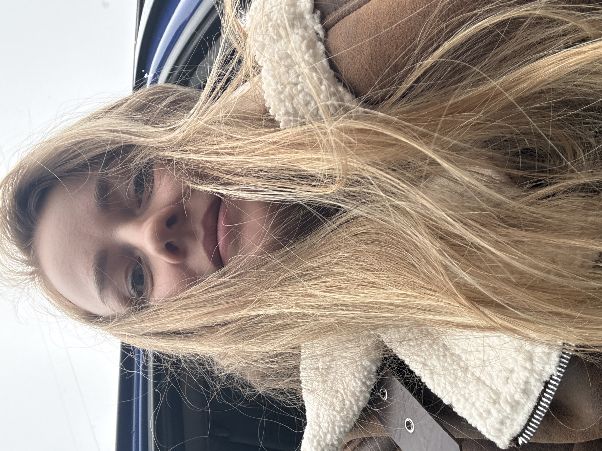 A fair-skinned woman with long, blonde hair partially obscuring her face is photographed in a selfie-style image. She is wearing a brown suede-like jacket with a white, fleece-lined collar. Part of a dark-colored car's exterior is visible in the upper left corner of the frame, suggesting she may be inside or near a vehicle. No specific features that would definitively pinpoint the location as Chaikovsky, Russia, are visible in the image.