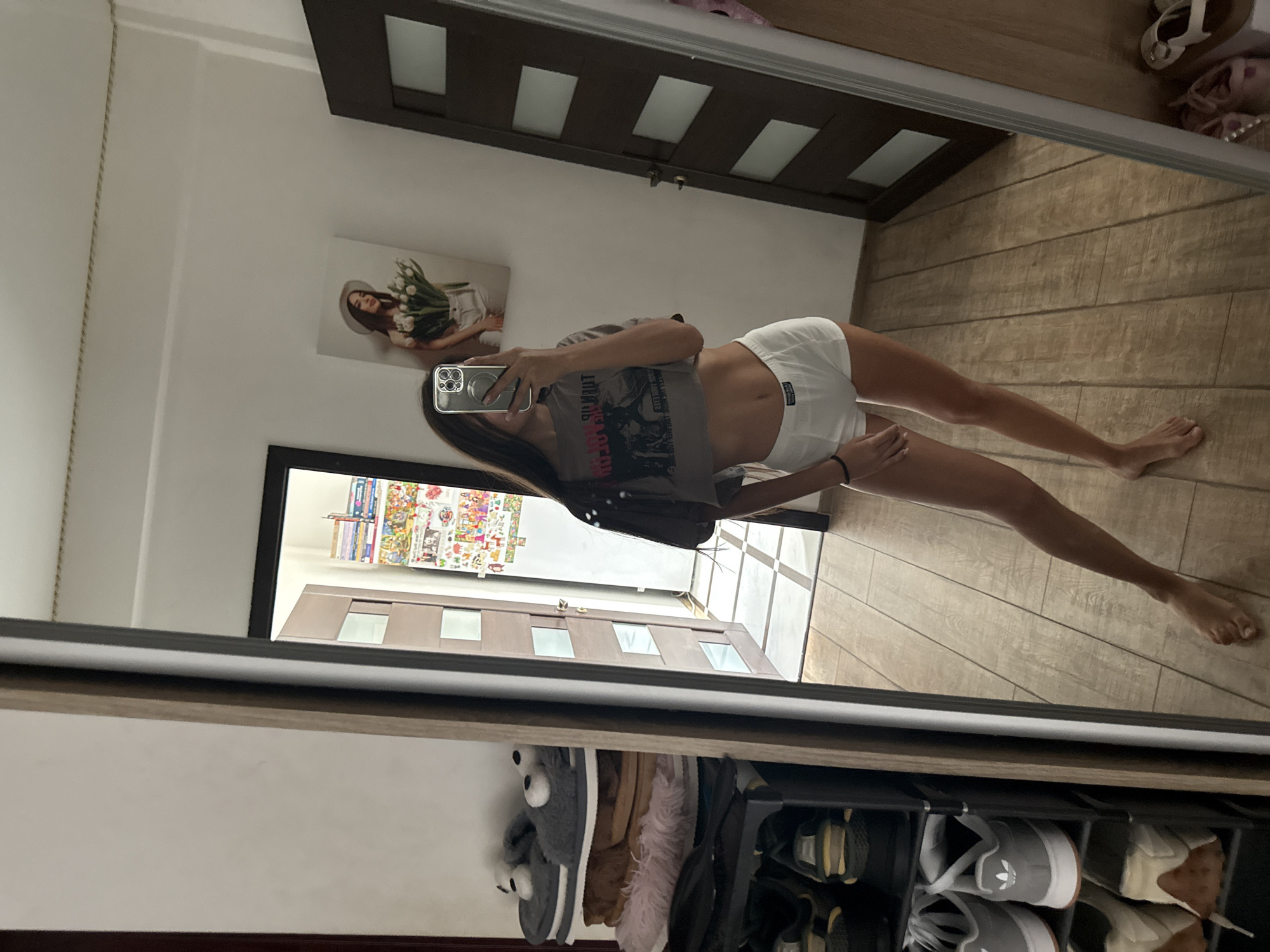 A young woman is standing in front of a mirror in a room. She is wearing a cropped t-shirt and white shorts. She is holding a phone and taking a selfie. In the reflection, we can see a shoe rack with several pairs of shoes and slippers, a door, and a painting on the wall. The room appears to be a bedroom or a dressing room.