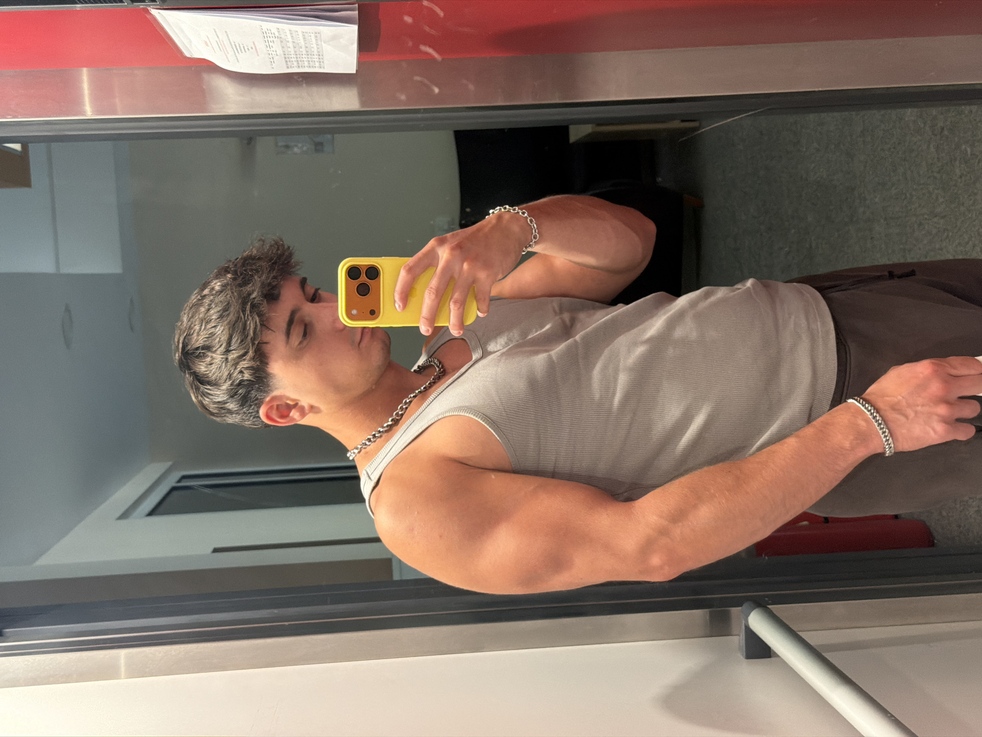 A young man with dark, tousled hair takes a mirror selfie in what appears to be an elevator. He is wearing a light gray ribbed tank top, brown sweatpants, and silver chain necklaces and bracelets. He is holding a bright yellow smartphone in his right hand, angled to capture his reflection. The elevator interior is visible, with metallic walls and a gray floor. A red panel is on the right side of the elevator, with a white document clipped to it. A metal handrail is visible on the left. The overall lighting suggests an indoor, possibly dimly lit environment. The specific location in Sofia, Bulgaria, cannot be determined from the visual information provided.