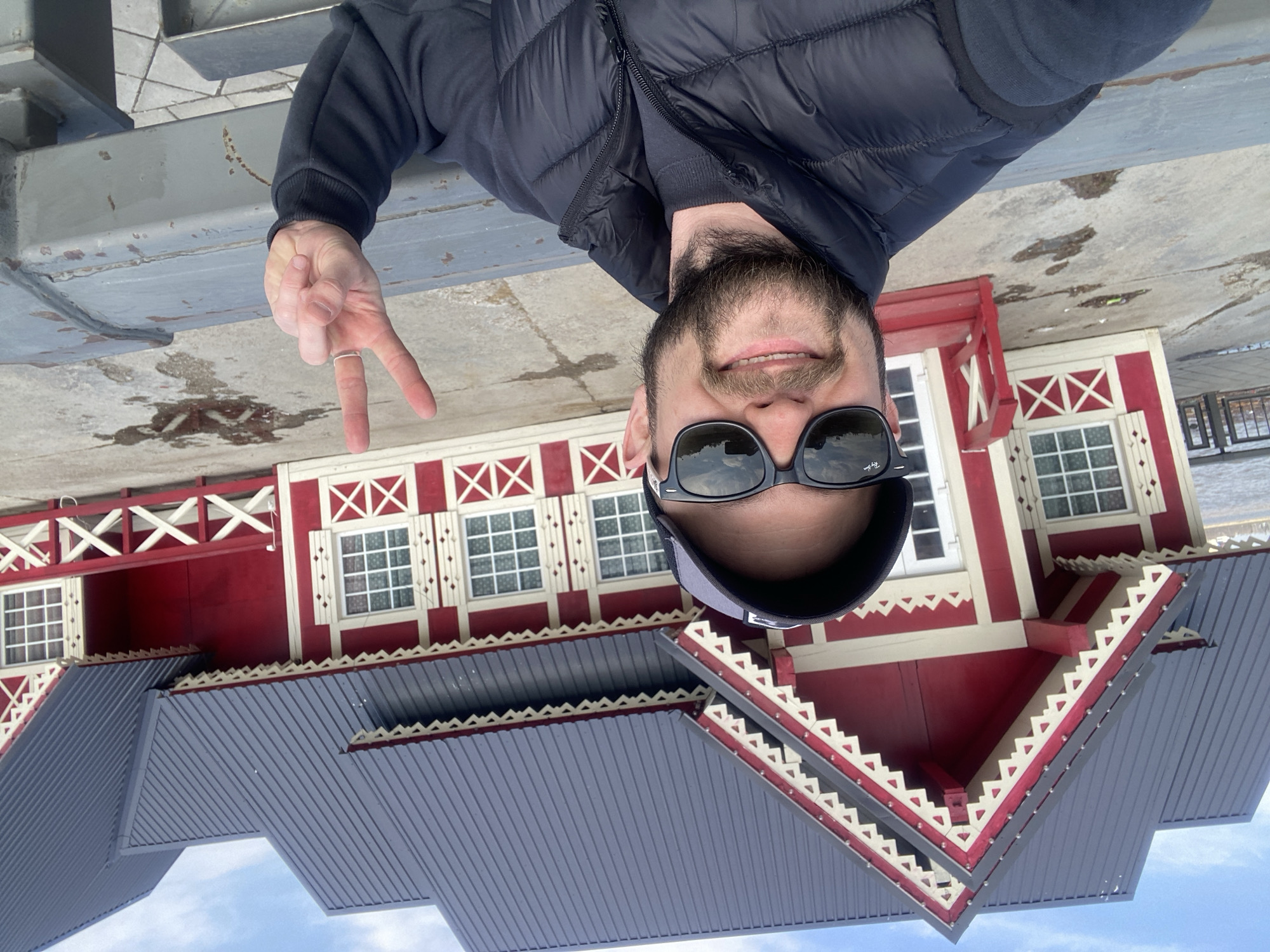A man is standing and taking a selfie. He is wearing sunglasses, a dark jacket, and a hat. He is making a peace sign with his left hand. Behind him is a red and white building with a gray roof. Based on the architecture, the building is likely located in Kazan, Russia.