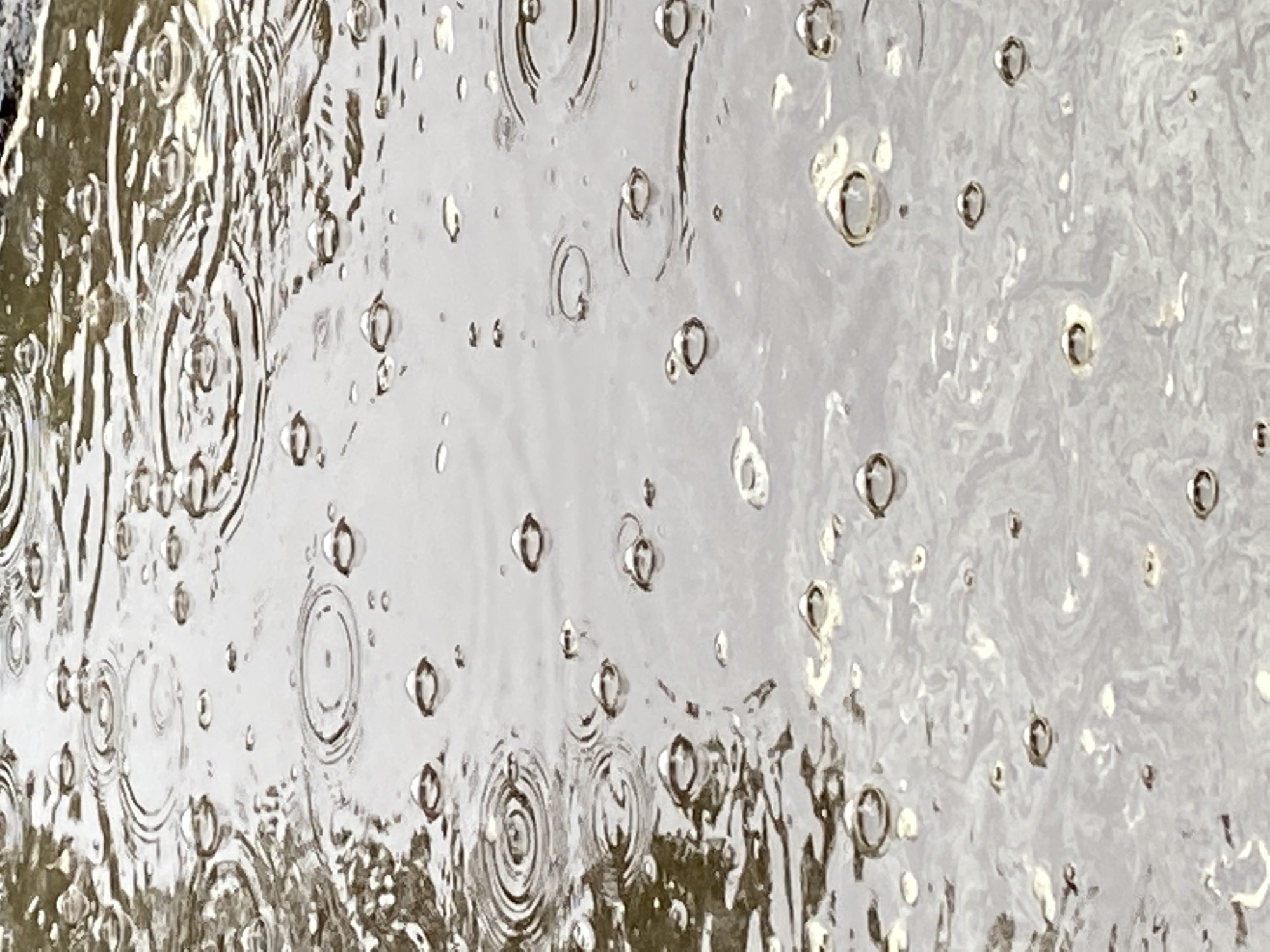 The image shows a close-up view of a surface covered in water, possibly a puddle or wet pavement. Raindrops are creating ripples and circular patterns on the water's surface. Some raindrops are visible as small, clear bubbles. The background reflects a blurred, muted color, suggesting an urban environment. There is no indication of the location being Chaikovsky, Russia, or any specific location. No people are visible in the image.