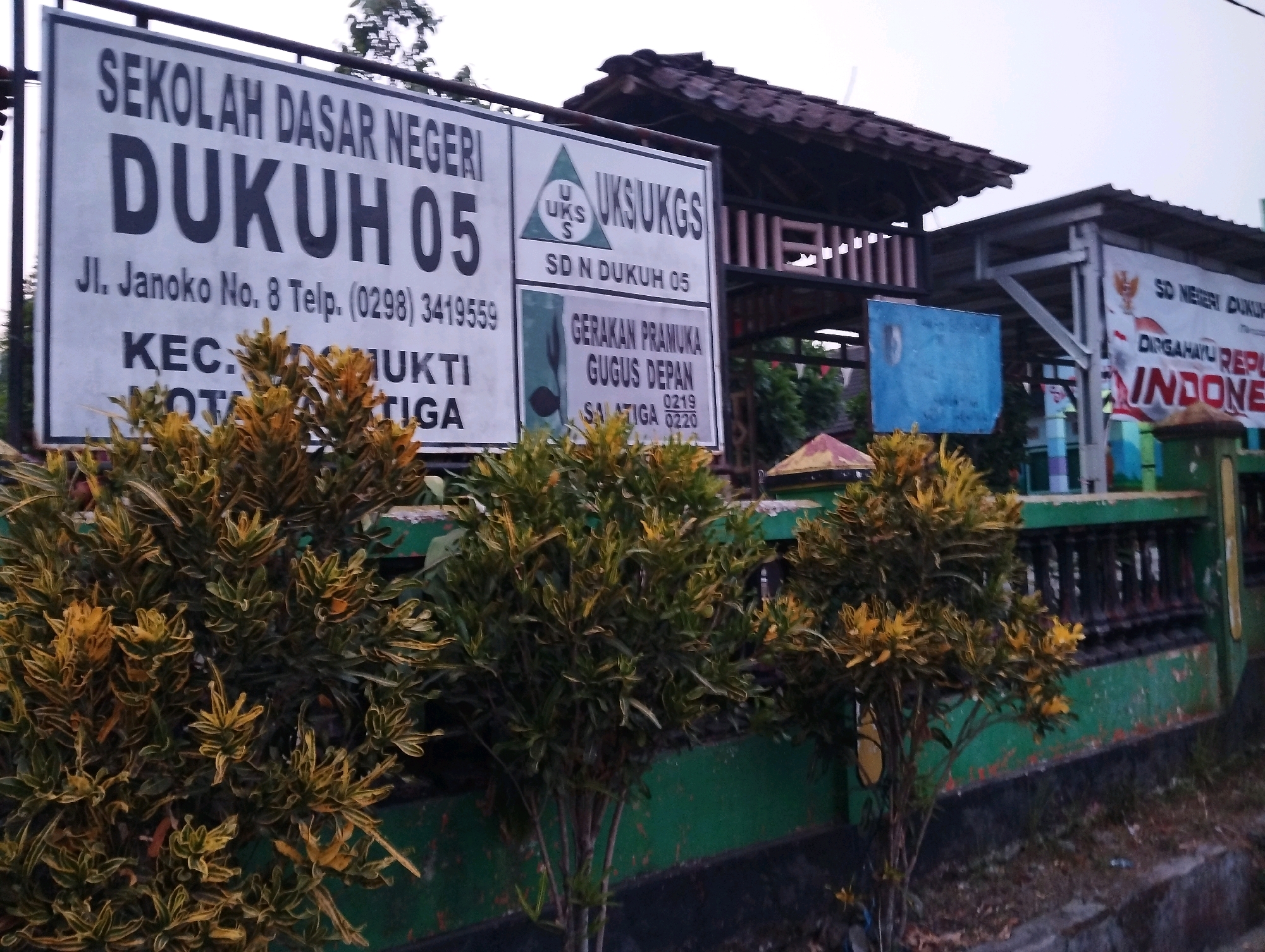 The image shows the entrance to a school in Salatiga, Indonesia. A large white sign with black lettering reads "SEKOLAH DASAR NEGERI DUKUH 05". Below this, it states "JL. Janoko No. 8 Telp. (0298) 3419559". Further text on the sign, partially obscured by foliage, indicates "KEC. MUKTI" and "KOTA TIGA".
To the right of the main sign is another sign with "UKS UKS" within a green triangle emblem, and below it "SD N DUKUH 05". Below this section is a sign for "GERAKAN PRAMUKA GUGUS DEPAN" with additional text "SALATIGA 0219 0220".
In front of the signs, decorative plants with variegated green and yellow leaves are visible. Behind the signs, there are structures with tiled roofs and some buildings that appear to be part of the school. A banner with red and white lettering, partially visible, reads "INDONESIA" and appears to have the Garuda Pancasila emblem, suggesting a national holiday or celebration. The environment appears to be outdoors, with some overgrown vegetation and a concrete walkway. The sky is overcast.