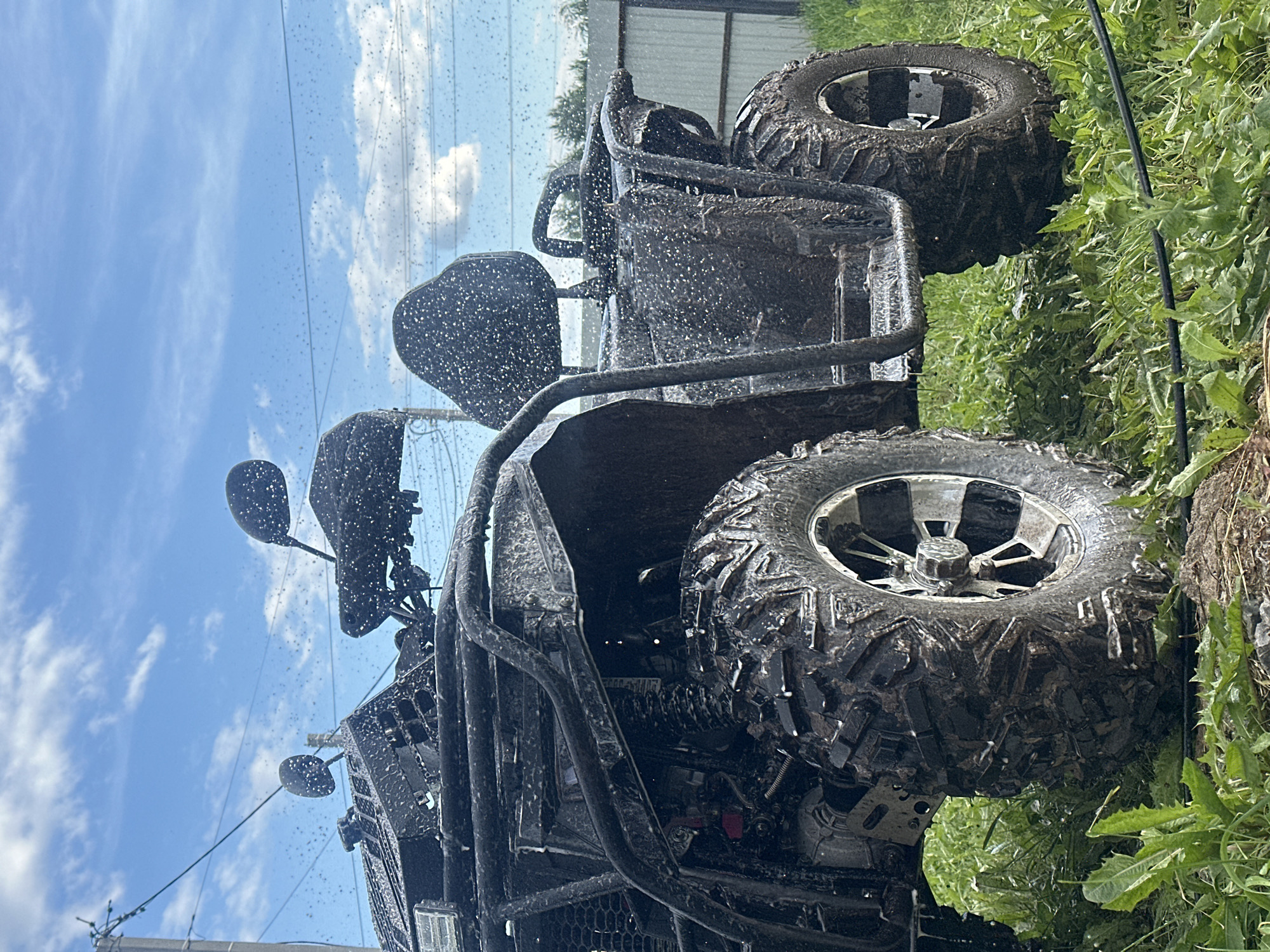 An all-terrain vehicle (ATV) is depicted stationary on a patch of green grass and dirt, partially covered in mud, located in Kharnavy, Russia. The dark-colored ATV is thoroughly coated with numerous small water droplets, indicating it is either undergoing a wash or has recently been exposed to a significant amount of water. Its knobby, muddy tires feature multi-spoke metallic rims, with the brand "WANDA" visible on the front-right tire. Structural elements such as a protective metal frame, suspension components, a dark seat, and two side mirrors are also visible and covered in water droplets. In the immediate foreground, vibrant green grass and disturbed soil are present, with a dark hose or cable partially obscured by the vegetation. The background on the left features a utility pole with multiple overhead power lines. To the right, a light-colored corrugated metal fence is visible, with trees extending beyond it. The scene is set under a bright blue sky with scattered white clouds. No individuals are visible in the image.