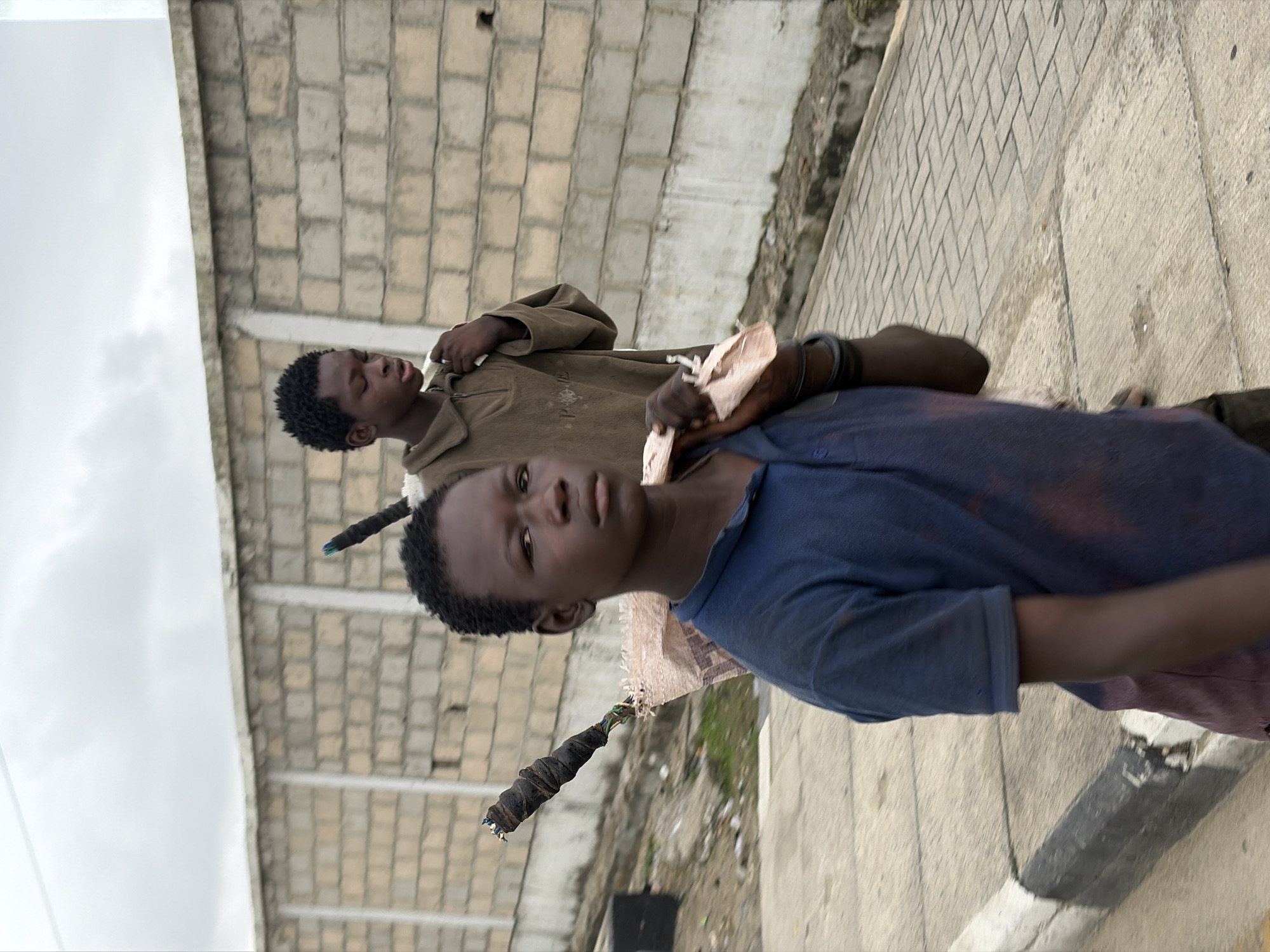Two young boys are walking on a paved sidewalk in front of a light-colored brick wall. The boy in the foreground, wearing a blue t-shirt and what appears to be a maroon garment underneath, is looking towards the camera. He is carrying a bundle wrapped in a light-colored sack over his left shoulder, supported by a long object that extends diagonally from behind him. The second boy, further back and to the right, is wearing a brown collared shirt and has his right hand on his chest. He is also carrying something over his shoulder. The environment appears to be an urban setting with a cloudy sky. The location is Igboefon, Nigeria.