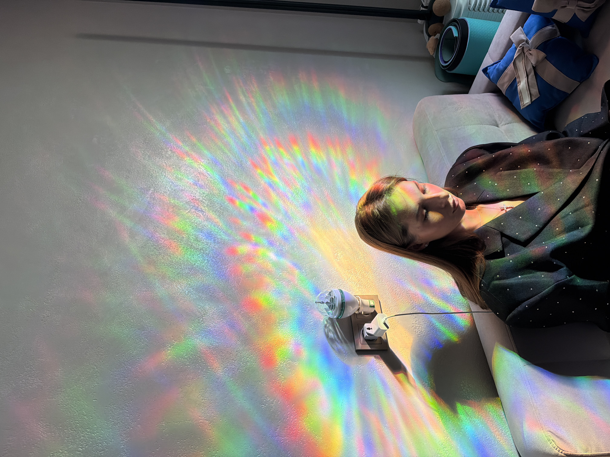 A woman is lying on a couch, her head resting on the armrest. She is wearing a dark blazer with small white dots. The room is illuminated by a rainbow-like light projection on the wall, casting colorful streaks across the surface. In the foreground, a power outlet is visible with two plugs, one of which appears to be connected to a small, faceted light fixture. Behind the woman, a blue decorative pillow with a beige bow and a rolled-up blue yoga mat are visible on the couch. The scene suggests a relaxed or contemplative moment, with the vibrant light creating an artistic and possibly ethereal atmosphere. The time of day is not specified, but the strong lighting suggests it could be daytime, with sunlight or artificial light being refracted. The location is identified as Бараўлянскі сельскі Савет, Belarus. There is no visible text in the image.
