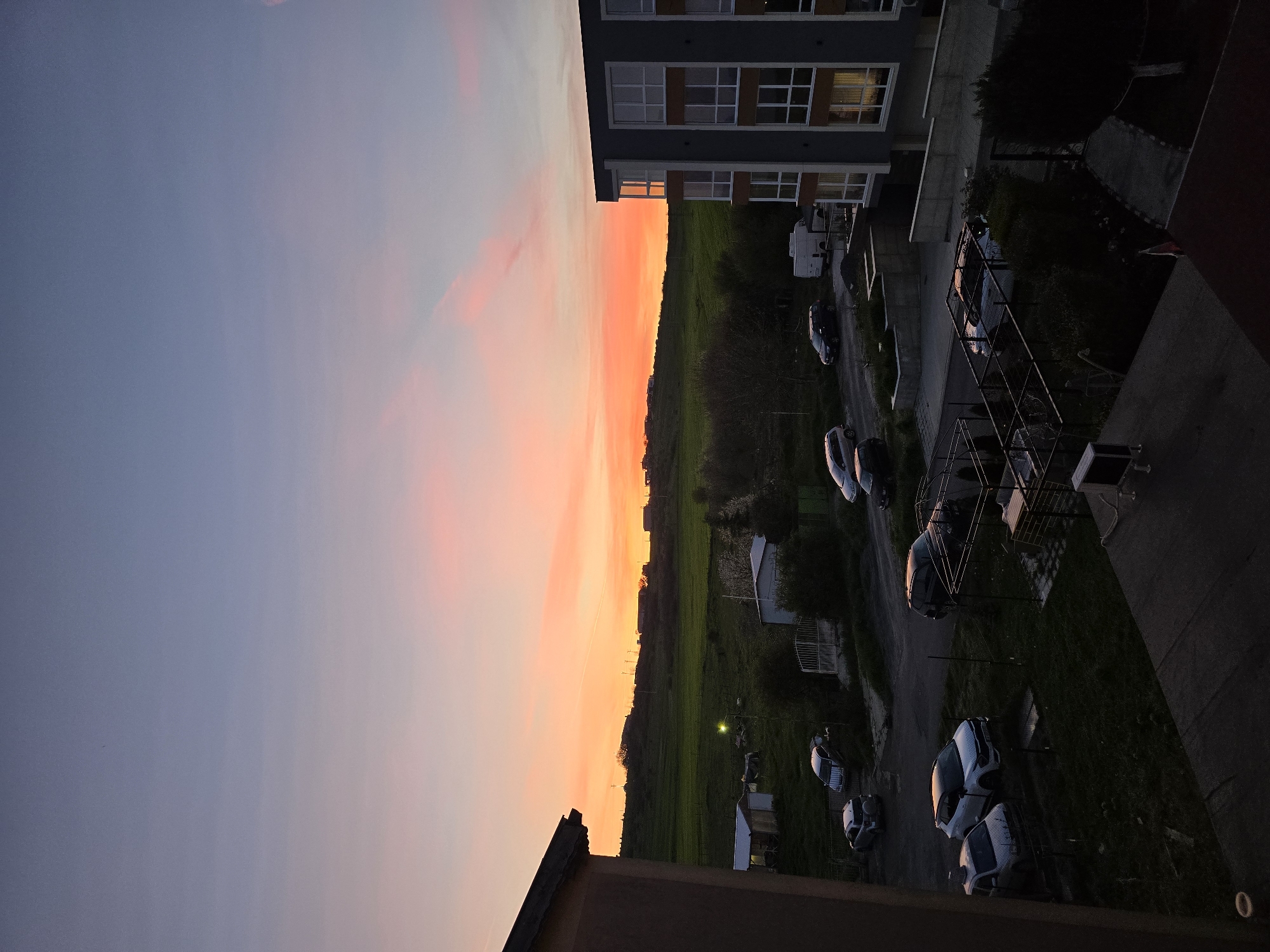 A slightly tilted, high-angle photograph captures an outdoor scene at either sunset or sunrise, with a vivid gradient of orange and pink in the sky. The sky transitions from light blue to a bright, warm orange near the horizon. Below the horizon, there's a landscape of fields, some of which appear green, and a line of trees. Buildings of varying sizes are dotted across the landscape.
To the right of the landscape, a gray apartment building with windows and dark window frames stands tall. Next to the building, a parking lot is visible, containing several cars. Some of the cars appear to be covered, possibly with car covers. A structure resembling a fence is also present.
The photo is taken from what seems to be a balcony or high vantage point, as a portion of a building's exterior is visible in the foreground. No people are visible in the frame. The lighting suggests dusk or dawn, casting long shadows.
The location is Burgas, Bulgaria.