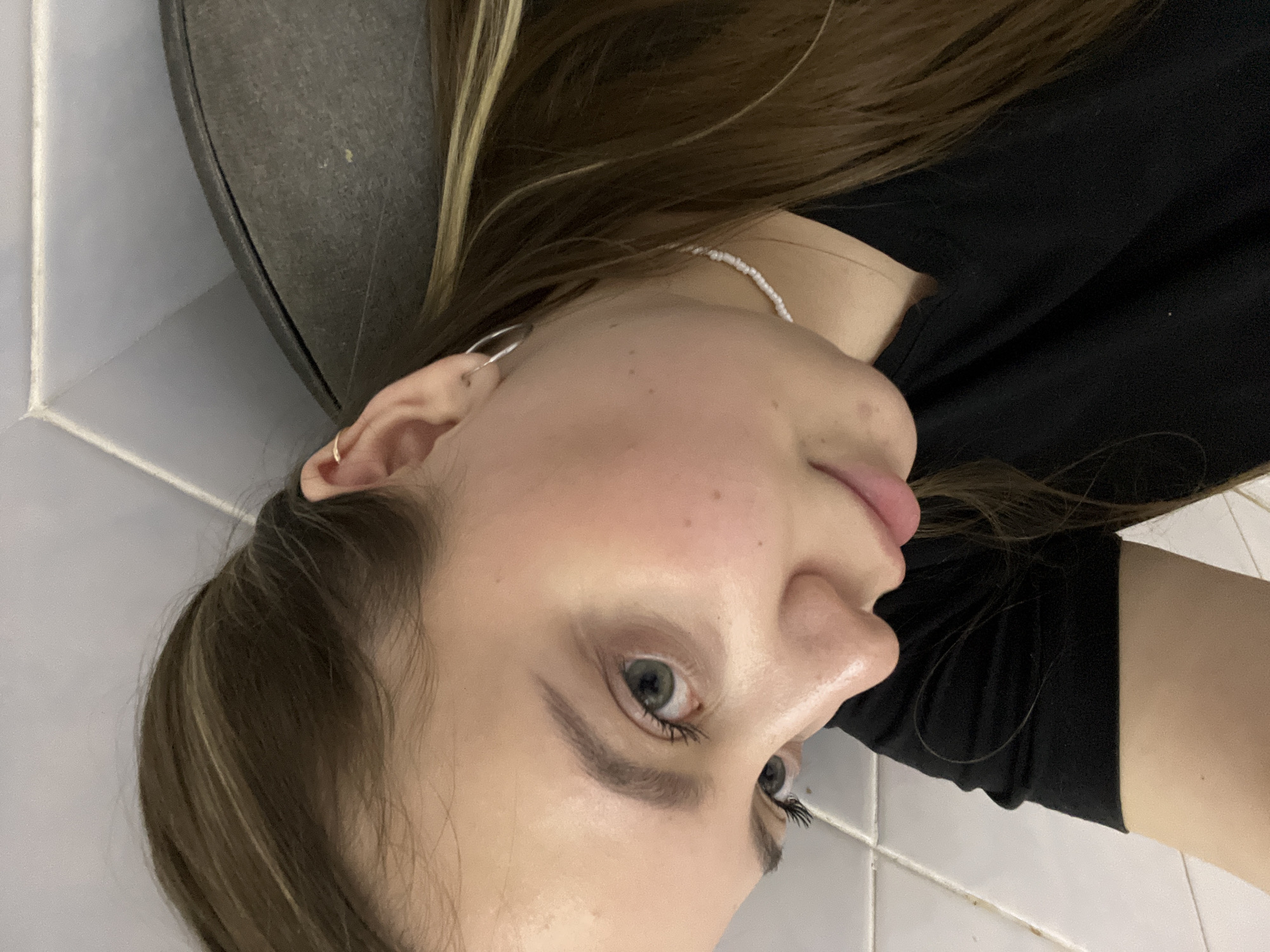 A close-up, tilted image shows a young woman with light skin, brown hair with blonde highlights, and grey-green eyes. She is wearing a black shirt, a pearl necklace, and hoop earrings. Her face shows slight acne scarring. The background includes a gray object, possibly a trash can, and white tiled walls. The location in Novopokrovskaya, Russia is unknown.