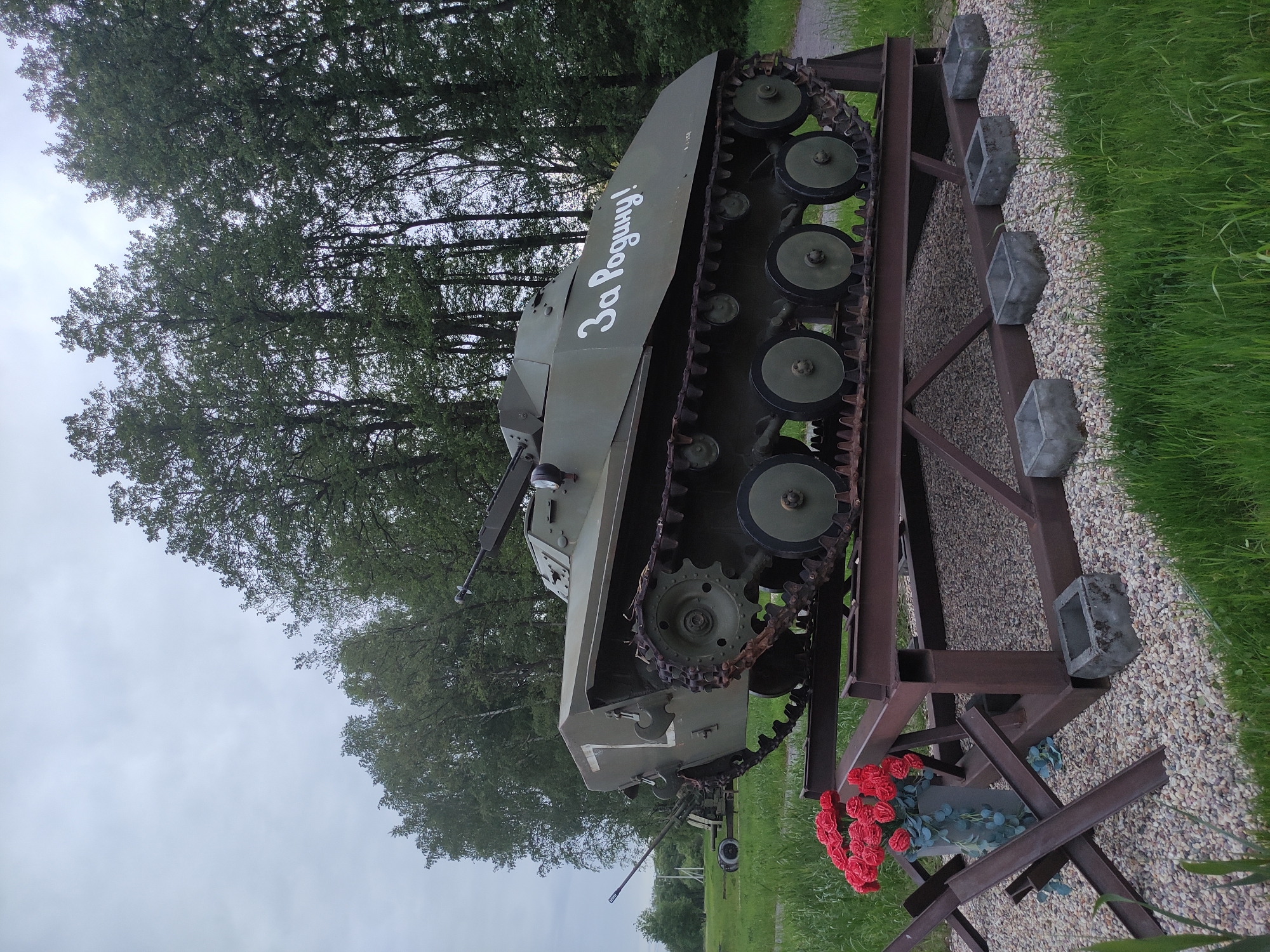 The image depicts a T-70 light tank, a World War II Soviet tank, mounted on a metal frame. The tank is painted olive green and has the inscription "За Родину!" ("For the Motherland!") in white on its side. The tank is located in Shevelkino, Russia. The background includes trees and greenery, suggesting a park or memorial site. At the base of the tank stand are red flowers. The tank is elevated on a dark metal frame. There are gray cube-shaped fixtures alongside the tank. The ground appears to be covered in light-colored gravel. A barrier made of metal is positioned in front of the flowers.